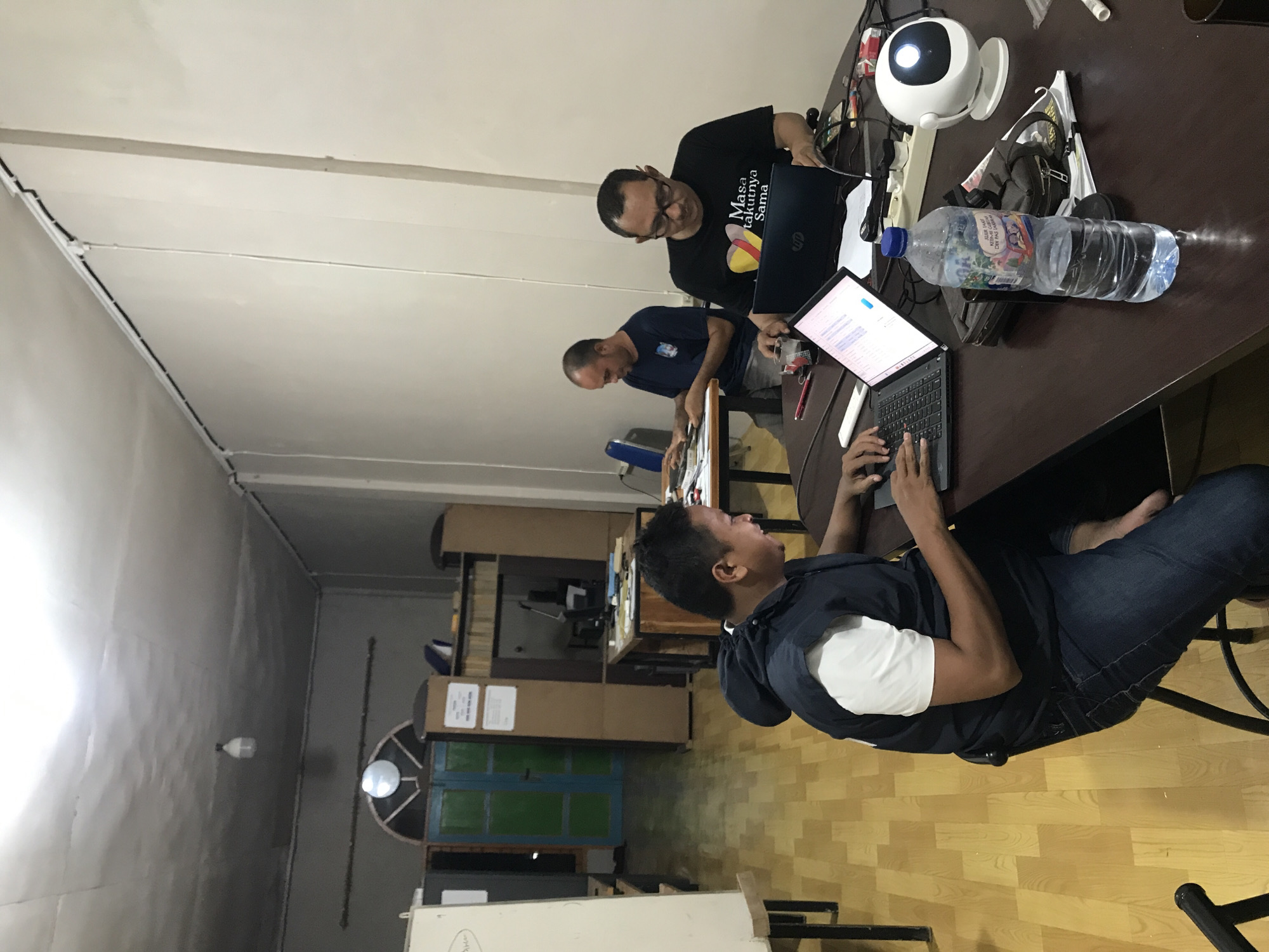 The image shows an indoor scene. Three men are present. Two are seated at a dark brown table using laptops. The man in the foreground is in a dark-colored shirt, jeans, and white undershirt. The man in the center is bald and wears a blue shirt. The man further in the back wears a black T-shirt with writing, and glasses, also using a laptop. On the table near these men is a white, circular object that looks like a camera. There is a bottle of water, and pens.
In the background, there are light gray walls and parquet flooring. A cupboard with green and blue doors is visible. There are miscellaneous boxes and other furniture. This is located in Tingkeummanyang, Indonesia.