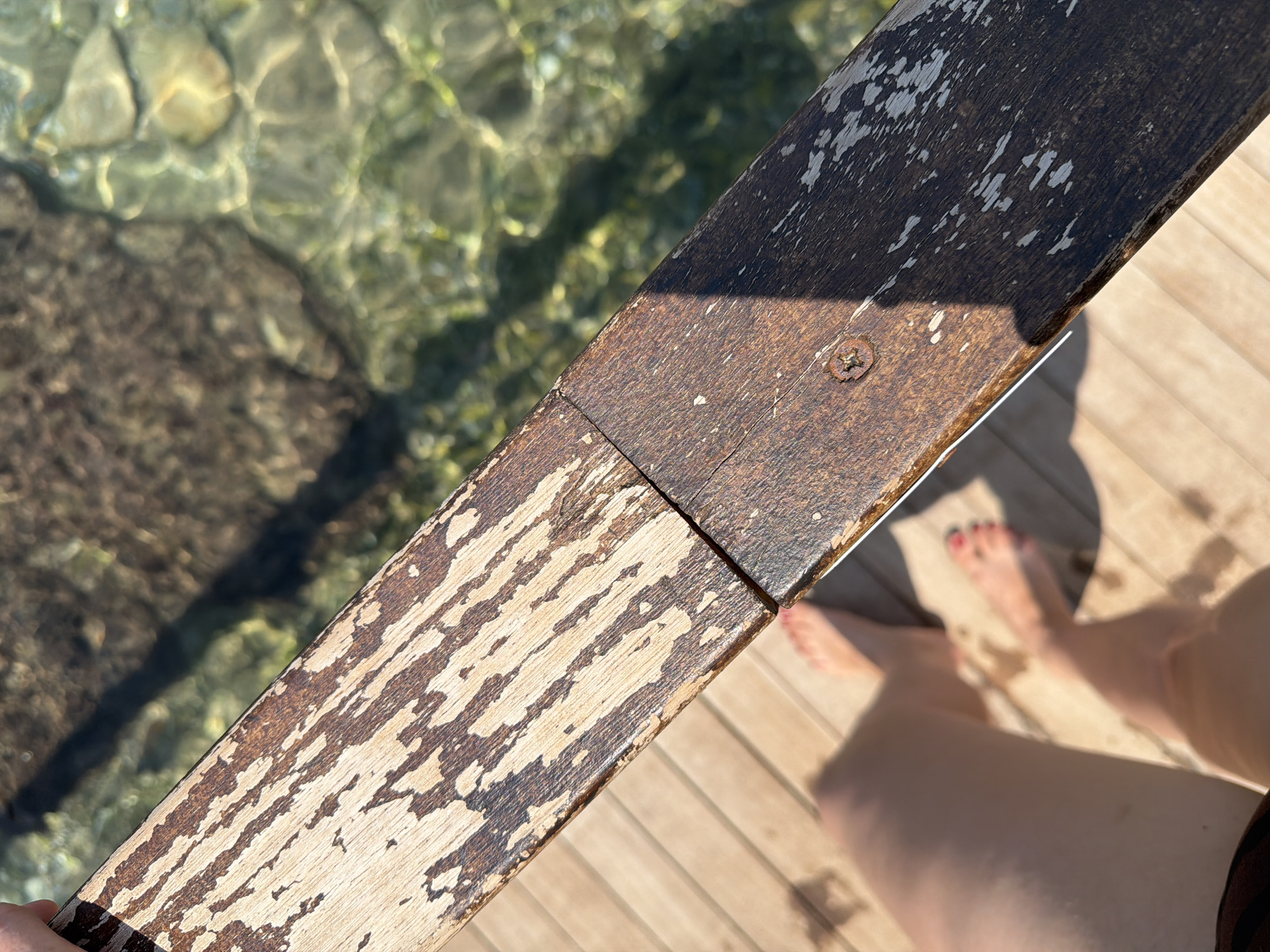 This is a close-up, high-angle shot of a weathered wooden railing over clear, shallow water. The railing, composed of two joined pieces of wood, shows significant wear, with areas of dark brown wood and lighter, faded patches where the finish has peeled. A single screw is visible on the lower right section of the railing.
Below the railing, a person's legs and feet are visible, standing on a wooden deck. The person appears to be barefoot, with red nail polish on their toes. The deck boards are light brown and appear to be wet in places, with small watermarks.
In the background, the clear water reveals the rocky bottom and some submerged plant life. The sunlight creates dappled patterns on the water's surface.
The overall scene suggests a location by the sea, possibly a pier or a boardwalk. Given the context of Antalya, Turkey, this could be a waterfront area like Konyaaltı Beach or a promenade along the Mediterranean coast. The weathered railing and direct sunlight suggest a warm, sunny day.