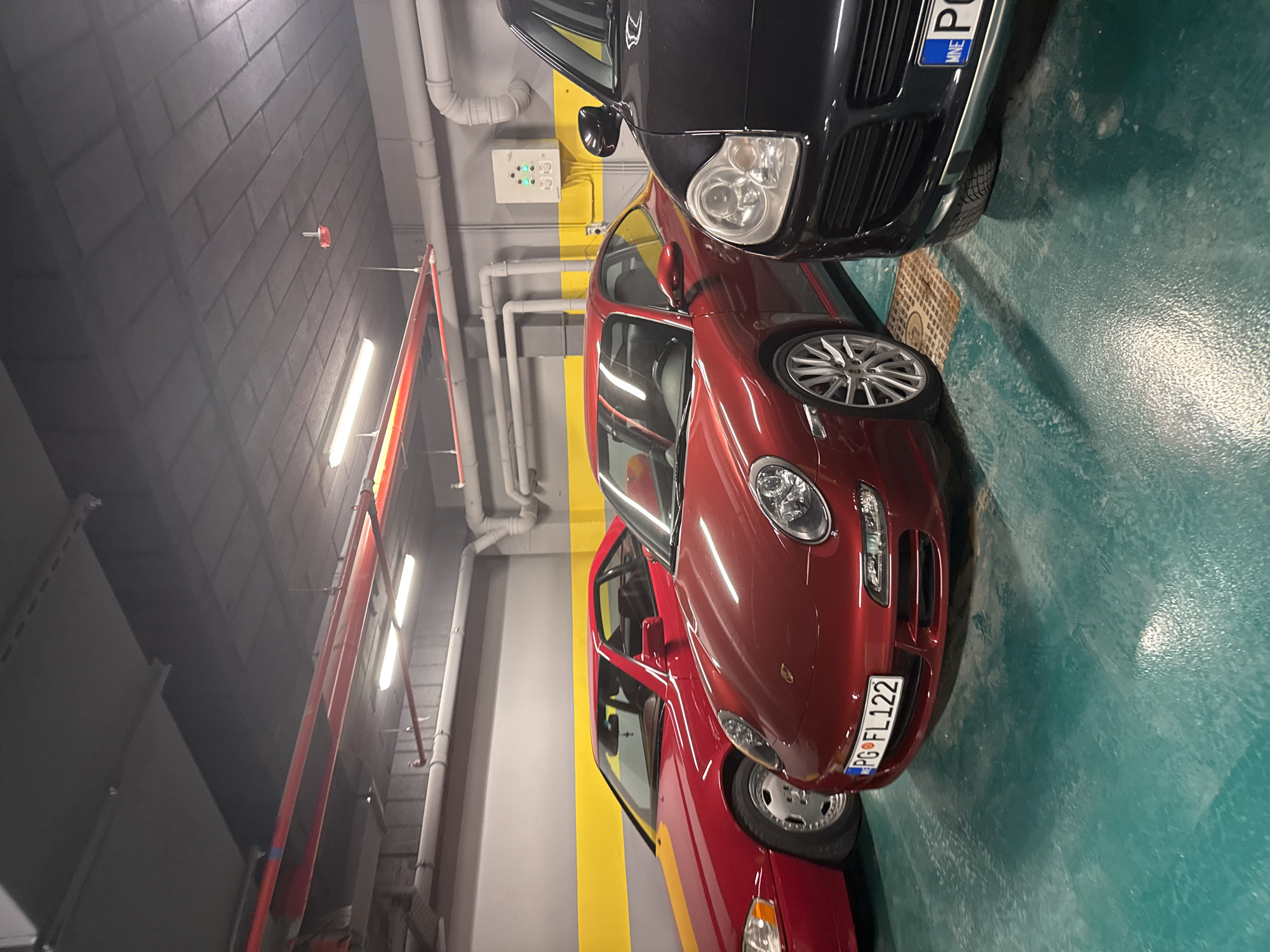 The image shows a section of an indoor parking garage. There are three cars visible. The closest car to the camera is a red Porsche, with a license plate that reads "PG FL122". Behind the Porsche is another red car. Next to the Porsche is a black SUV-type vehicle with a license plate beginning "MNE". The parking garage floor is painted in green, with yellow stripes on the walls indicating parking spots. Pipes and utility infrastructure are visible on the ceiling and walls of the garage. The location is not explicitly identified as Katun Bačko brdo, Montenegro, but the license plate "PG" is a likely indicator that the cars are registered in Montenegro. The image lacks any visible people, subjects, actions or interactions.