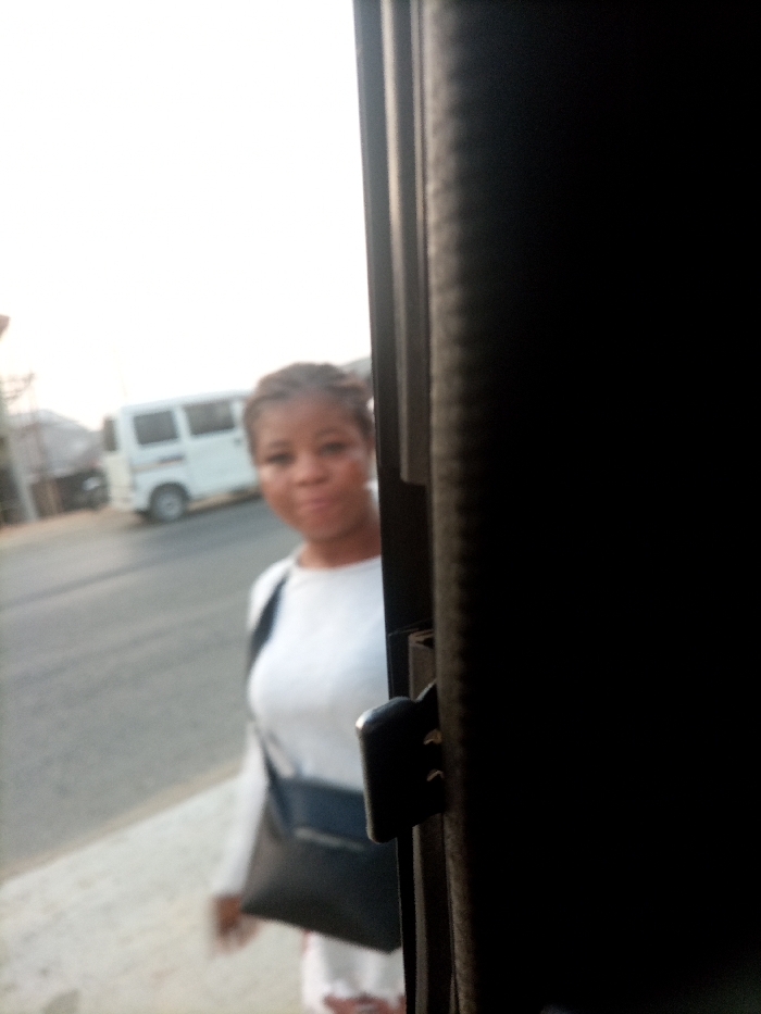 The image displays an outdoor scene in the city of Obehie, Nigeria, viewed from an interior perspective through an opening. A dark vertical structure, featuring a black latch mechanism, frames the right side of the image. A female individual is positioned in the left portion of the frame, out of focus, wearing a white long-sleeved top and a black shoulder bag across her body. Her hair is braided, and her face is oriented towards the camera. In the blurred background, a white minivan is visible on a paved road. Distant buildings and structures are indistinct, beneath a bright, uniformly lit sky.