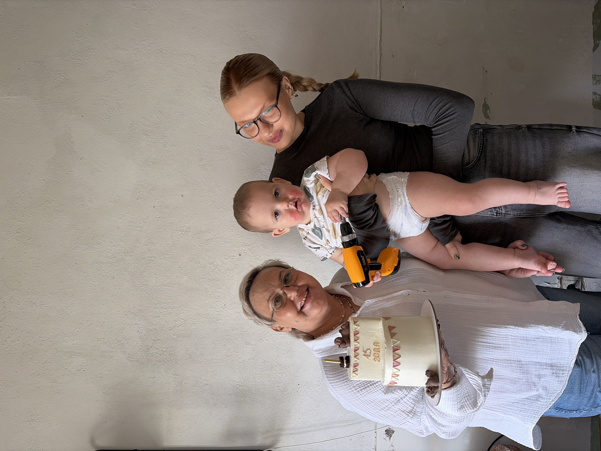 The image shows three people indoors. An older woman with short, light hair and glasses smiles while holding a white cake decorated with pink triangles and the number "45 2000". A younger woman with glasses and braided hair holds a baby in a diaper, who is holding a yellow and black toy drill. The background is a light-colored wall. The image was taken in the city of Chaikovsky, Russia.