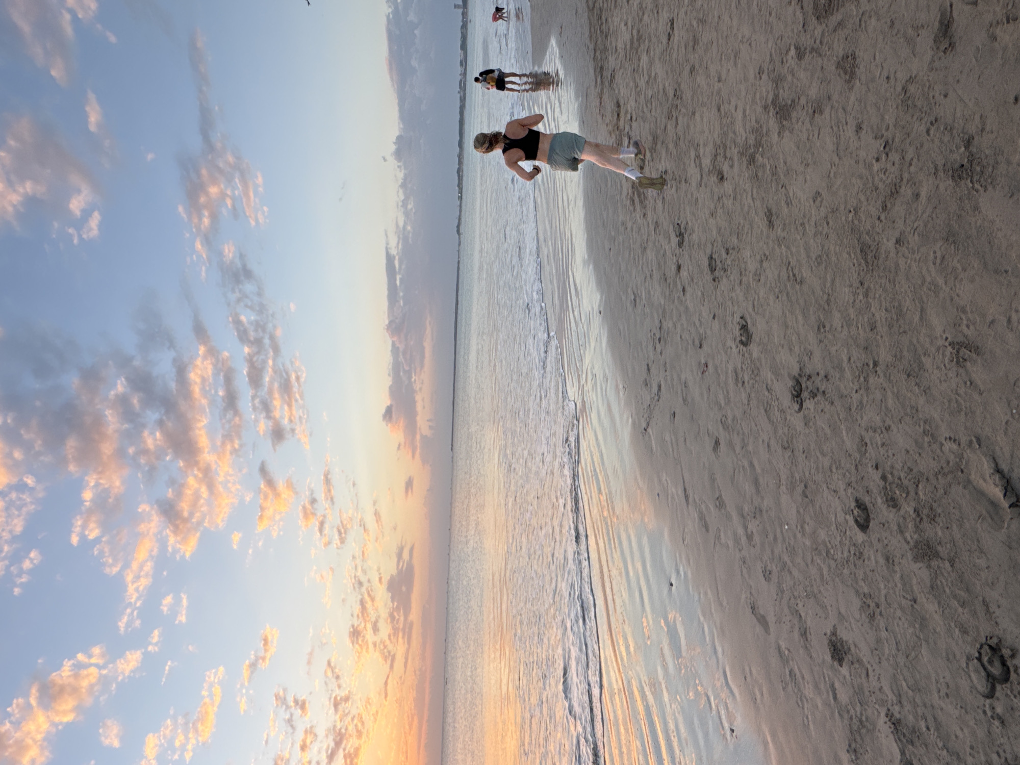 This is a vertical, eye-level, medium shot captured during twilight at a beach in Kuta Selatan, Indonesia. The sky is a gradient of pale blue and soft orange, with scattered clouds tinged with pink and yellow. The ocean is calm, with gentle waves lapping at the shore, reflecting the colors of the sky.
In the foreground, a person with blonde hair tied back is running along the wet sand, facing away from the camera and towards the right of the frame. They are wearing a black sports bra and light green shorts, with white socks and athletic shoes. Their body is positioned mid-stride, indicating movement.
Further back on the beach, a couple stands close together, also facing away from the camera. The person on the left appears to be taller and is wearing dark clothing, while the person on the right is wearing lighter-colored clothing. To the far right of the frame, a small child is visible, bent over as if playing in the sand.
The beach itself is composed of damp sand, marked with numerous footprints. The water's edge creates a reflective surface on the sand, mirroring the sky and distant elements. In the far distance, a faint horizon line suggests land and possibly buildings. The overall atmosphere is serene and peaceful, characteristic of a beach at sunset.