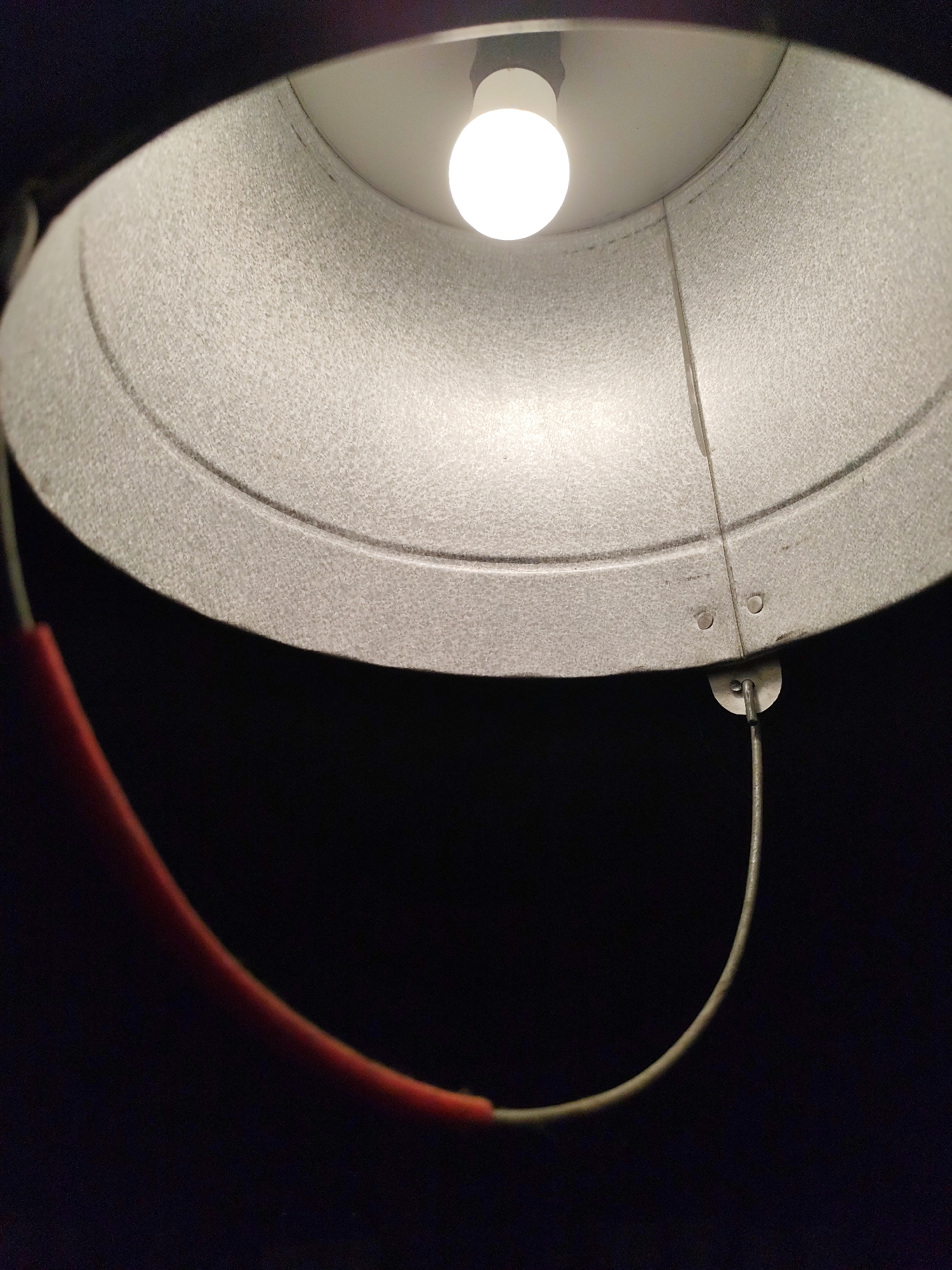 A low-angle shot captures the interior of a metal bucket. A bare incandescent lightbulb illuminates the bucket's interior. A metal wire handle, partially covered by a red sleeve on one side, is attached to the bucket. The background is dark, obscuring the setting. There is no evidence of this being located in Da Lat, Vietnam.