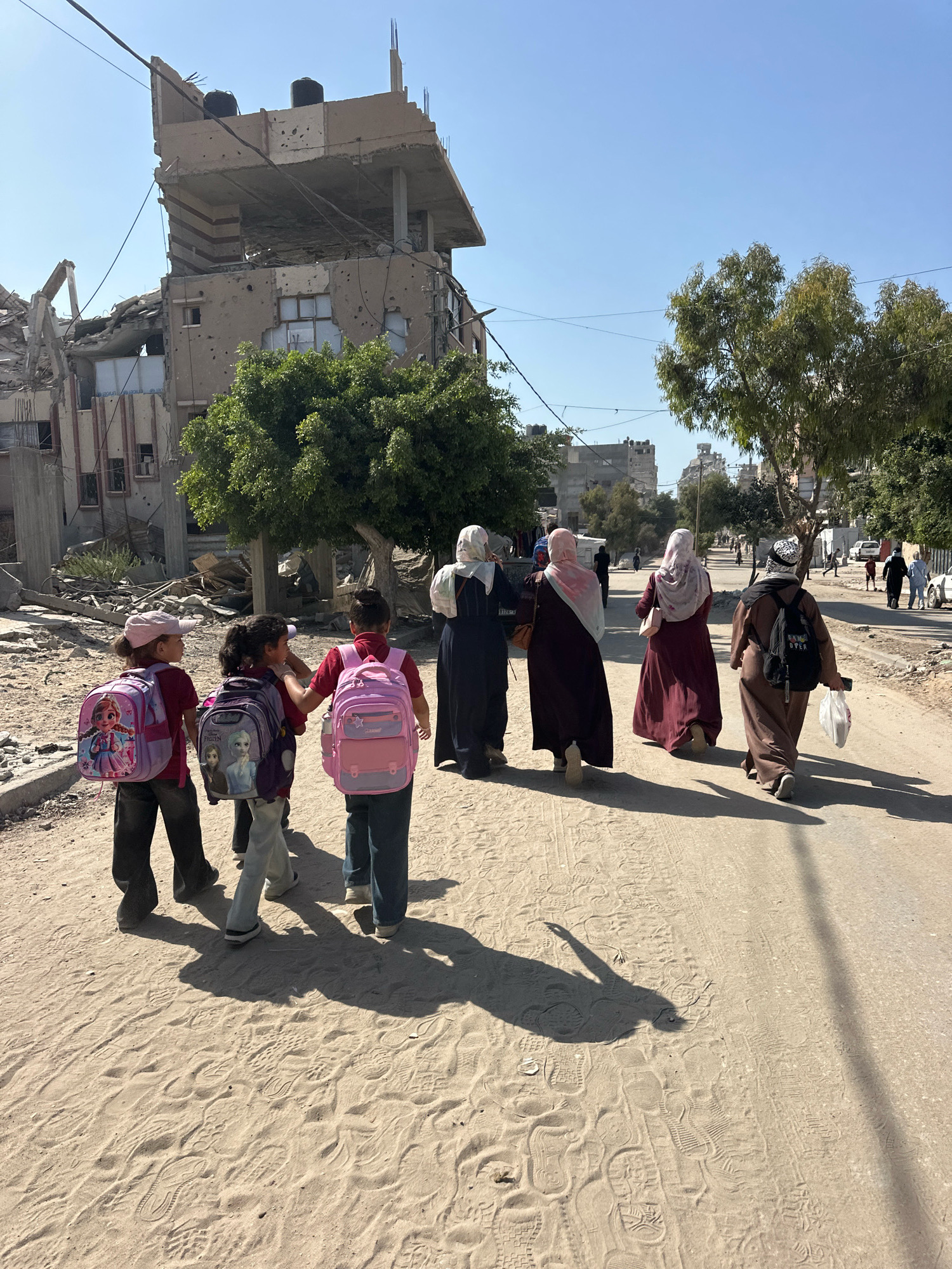In the city of Gaza, Palestinian Territories, a group of children and adults walk down a dusty road. In the background, a damaged building stands, displaying signs of structural compromise and bullet holes. The sky is clear and blue. Some of the children are wearing backpacks, suggesting they may be on their way to or from school. The adults are dressed in traditional attire, with some wearing headscarves. The overall scene depicts a stark contrast between the resilience of daily life and the visible impact of conflict in the environment.