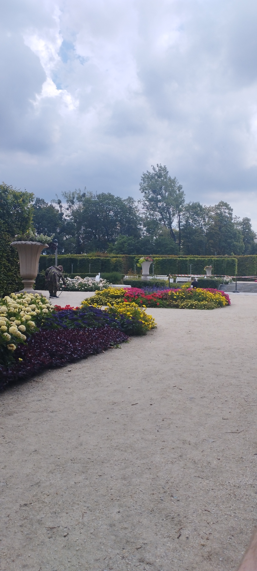 In this image, a person dressed in camouflage is tending to a vibrant flower bed in a park. The flower bed features a striking arrangement of yellow, red, pink, and purple flowers, bordered by dark purple foliage. To the left, a large, ornate vase stands beside a manicured green hedge. Behind the flowers, a hedge forms a backdrop, with more trees visible further in the distance. Several white benches are also present, suggesting a public space. The sky is overcast with dramatic clouds. The scene captures a moment of gardening or maintenance within a well-kept park setting.
