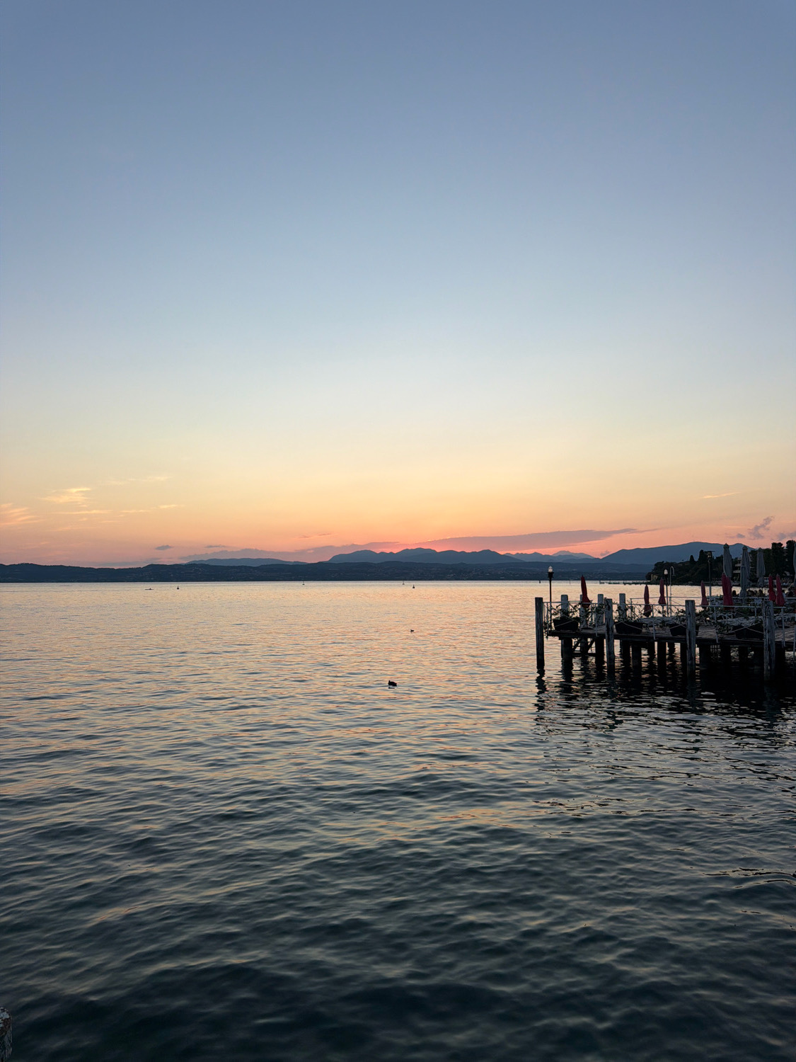 The image captures a serene sunset over Lake Garda in Sirmione, Italy. The sky transitions from a pale blue at the top to soft hues of orange and pink near the horizon, where a range of mountains is silhouetted against the fading light. The calm water reflects the sky's colors and is gently rippled. In the foreground, a wooden pier extends into the lake, featuring a few lampposts and a collection of tables and chairs, suggesting an outdoor seating area for a restaurant or cafe. There are no people visible in the scene, but a lone duck is present on the water.