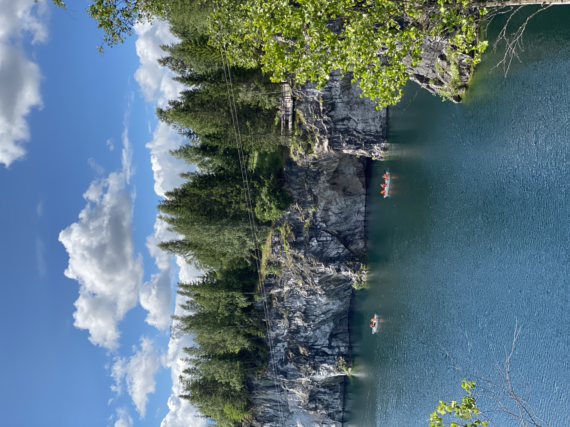 The image depicts a scenic view of the Ruskeala Mountain Park in Russia, a former marble quarry now a popular tourist destination. The scene features a deep, turquoise-blue lake surrounded by towering, sheer marble cliffs. Lush green trees and dense forest cover the tops of the cliffs, contrasting with the stark gray and white of the rock formations. The sky above is a vibrant blue, dotted with fluffy white clouds.
In the water, two small white boats are visible. The boat in the foreground has at least three people wearing orange life jackets, suggesting they are engaged in recreational activity on the lake. Another boat is further in the distance, also appearing to have occupants.
Along the cliff face, a wooden walkway or observation deck is present, with several people standing on it, likely admiring the view. A thin wire or cable stretches across the upper part of the image, possibly for a zipline or other attraction. The overall impression is one of natural beauty and outdoor recreation, with the geological formations of the quarry serving as a dramatic backdrop.