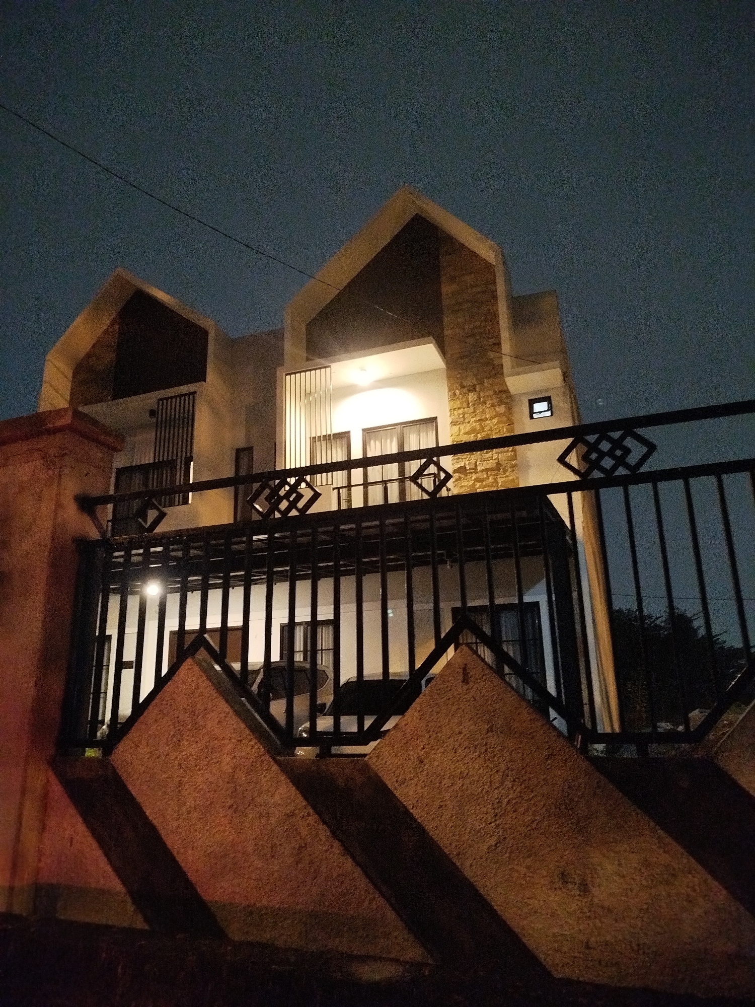 This is a low-angle, full-shot photograph taken at night, showing the exterior of a modern, multi-story house in Depok, Indonesia. The house features a unique architectural design with sharp, angular rooflines and is illuminated by interior lights. A black metal fence with geometric patterns runs across the foreground, partially obscuring the view of the house. The fence is supported by a reddish-brown concrete pillar on the left. Shadows are cast on the ground by the fence and the pillar, indicating an external light source. The sky is dark and clear, with a faint glow of light. A thin wire stretches diagonally across the upper portion of the frame. No people are visible in the image.