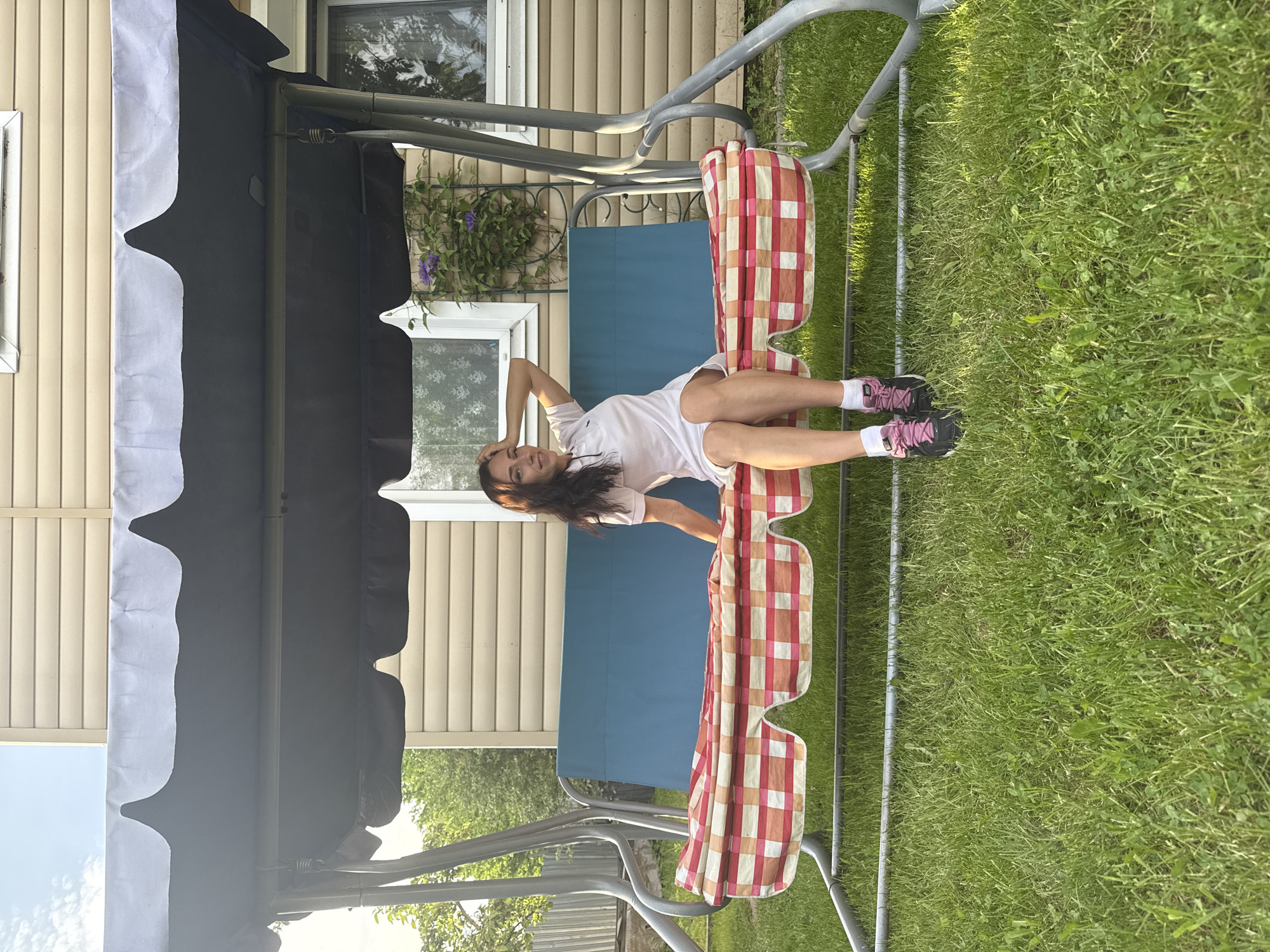 This image captures an outdoor scene in Saint Petersburg, Russia, featuring a young adult female seated on a swing set in a residential backyard. The woman, with long dark hair, wears a white polo-style dress, white socks, and black and pink Nike athletic shoes. Her left arm is raised, with her hand resting on the back of her head as she looks towards the camera with a slight smile. The swing set has a grey metal frame and a dark grey canopy with a white scalloped edge. The swing seat is blue, partially covered by a plaid cushion or blanket in shades of red, orange, and white. Behind the swing set, a house with beige horizontal siding and white-framed windows is visible. A climbing plant with purple flowers grows on a trellis against the house to the right. Green trees and foliage are present on the left side of the background, and a metal fence is partially visible. The foreground consists of vibrant green grass, suggesting a sunny outdoor environment.