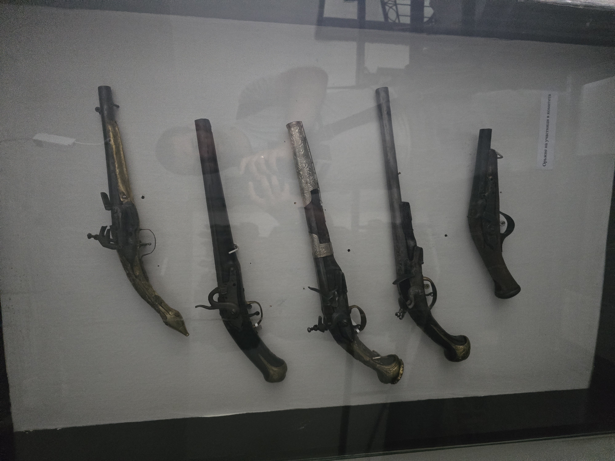 The image displays five antique flintlock pistols arranged horizontally inside a glass display case against a light grey background. The scene is located in a museum exhibit in Teteven, Bulgaria.
From top to bottom:
1. The topmost pistol features a long, dark barrel and a distinctive gold-colored handle and fore-stock, intricately shaped with what appears to be an animal head motif (possibly a serpent or mythical creature) forming the front part of the stock near the muzzle. The flintlock mechanism is visible.
2. The second pistol has a dark, plain wooden stock and a dark barrel. It appears less ornate than the first. Its flintlock mechanism is also visible.
3. The third pistol from the top features a dark wooden stock with a prominent, ornate gold-colored pommel or butt cap. The barrel section near the stock is adorned with a broad, silver-colored band featuring intricate engraved scrollwork. The flintlock mechanism is present.
4. The fourth pistol closely resembles the third, with a dark wooden stock and a gold-colored, ornate pommel. The barrel is dark, and its flintlock mechanism is visible.
5. The bottommost pistol is shorter and appears simpler in design, with a dark, less decorated stock and barrel.
Reflected in the glass display case, a person (likely the photographer) is visible holding a mobile phone or camera, primarily in the middle and lower-middle sections of the image. The person appears to be wearing a light-colored shirt or jacket. Shadows from the surrounding environment, including what might be a window frame or display structure, are also visible in the reflections on the right side.
In the bottom right corner of the display case, a white label with black Cyrillic text is affixed. The text reads "Оръжие на участници в комитета," which translates to "Weapons of committee members." This suggests the pistols are historically significant artifacts related to revolutionary or national liberation committees, consistent with the historical context of Teteven, Bulgaria.