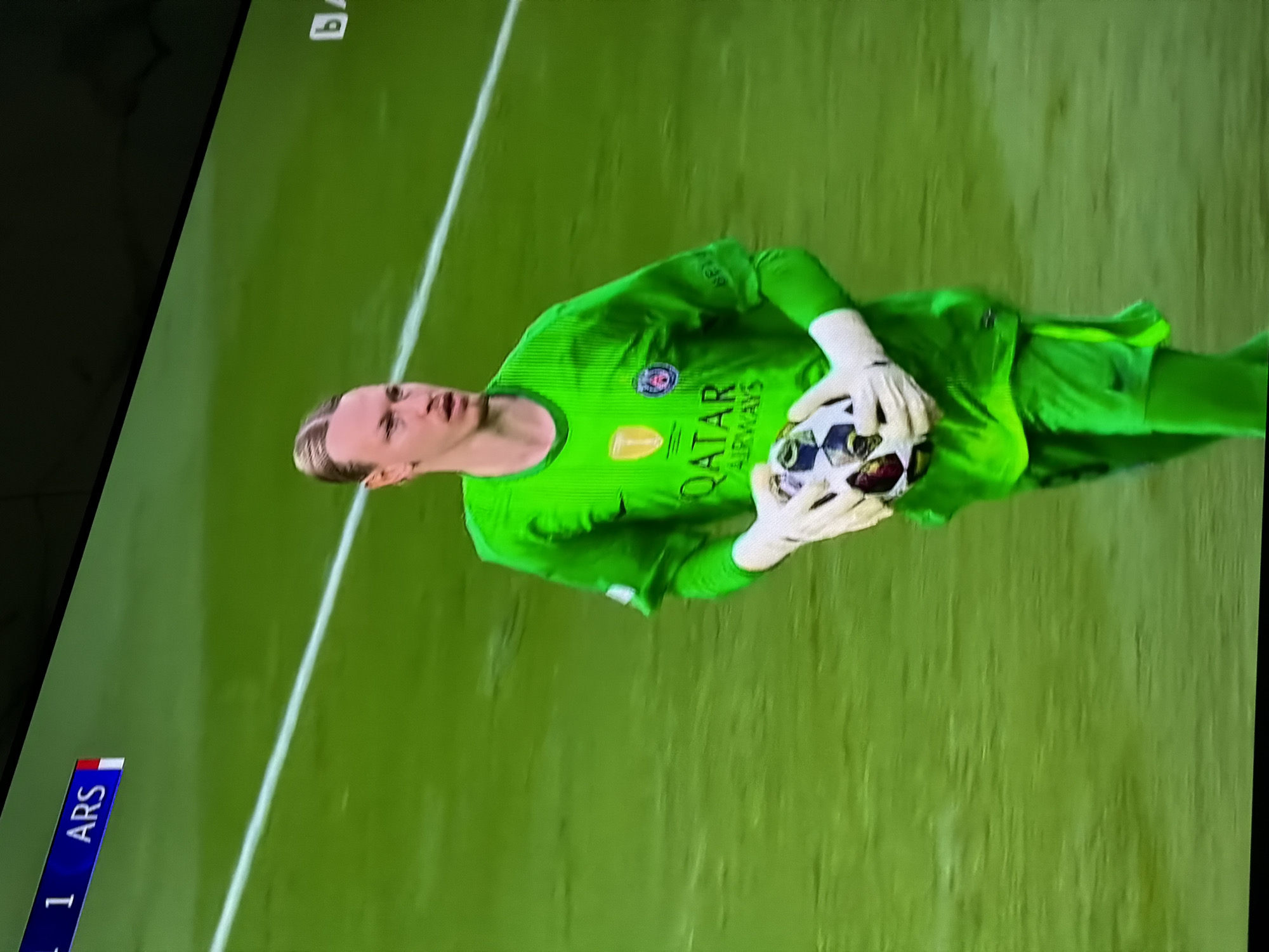 The image shows a soccer game. The goalkeeper, wearing a bright green uniform with "QATAR AIRWAYS" on the chest, is holding a soccer ball. The field is green. The score is visible, showing "1" for "ARS" (Arsenal). There is no visible evidence to suggest the location is Asenovgrad, Bulgaria.