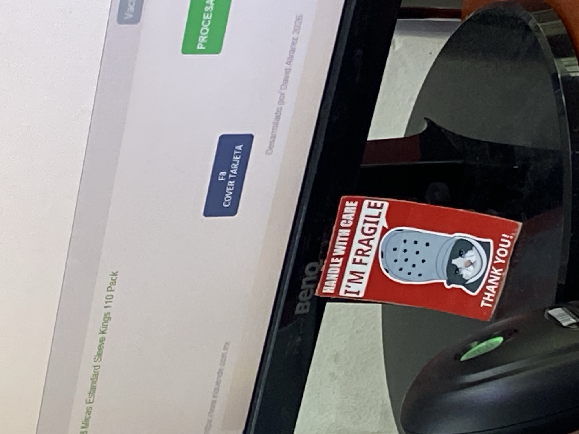 The image shows a BenQ computer monitor with a sticker on its side displaying a cat inside a Croc shoe, accompanied by the phrases "HANDLE WITH CARE", "I'M FRAGILE" and "THANK YOU!". The screen displays a webpage in Spanish, possibly related to product sales ("Micas Estandard Sleeve Kings 110 Pack"). Two buttons labeled "Vac" and "PROCESA" and another label named "COVER TARJETA" can be seen on the webpage as well. The text "Desarrollado por David Alvarez 2026" also appears on the monitor screen. The monitor is placed next to a black scanner. The text "mps www.elduende.com.mx" is present at the bottom of the screen, which indicates that the photo may have been taken in Mexico City, Mexico.
