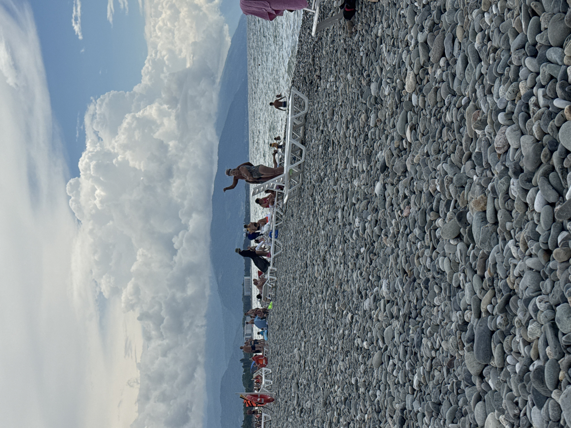 This image depicts a busy pebble beach scene in Sirius, Russia. The foreground is dominated by a vast expanse of smooth grey pebbles. In the midground, numerous people are spread across the beach, engaged in various activities. Several individuals are relaxing on white lounge chairs, while others are standing or walking. The sea is visible behind the beach, with gentle waves lapping at the shore. A line of mountains stretches across the horizon, partially obscured by clouds. The sky above is a mix of blue patches and large, fluffy white clouds, indicating partly cloudy weather.