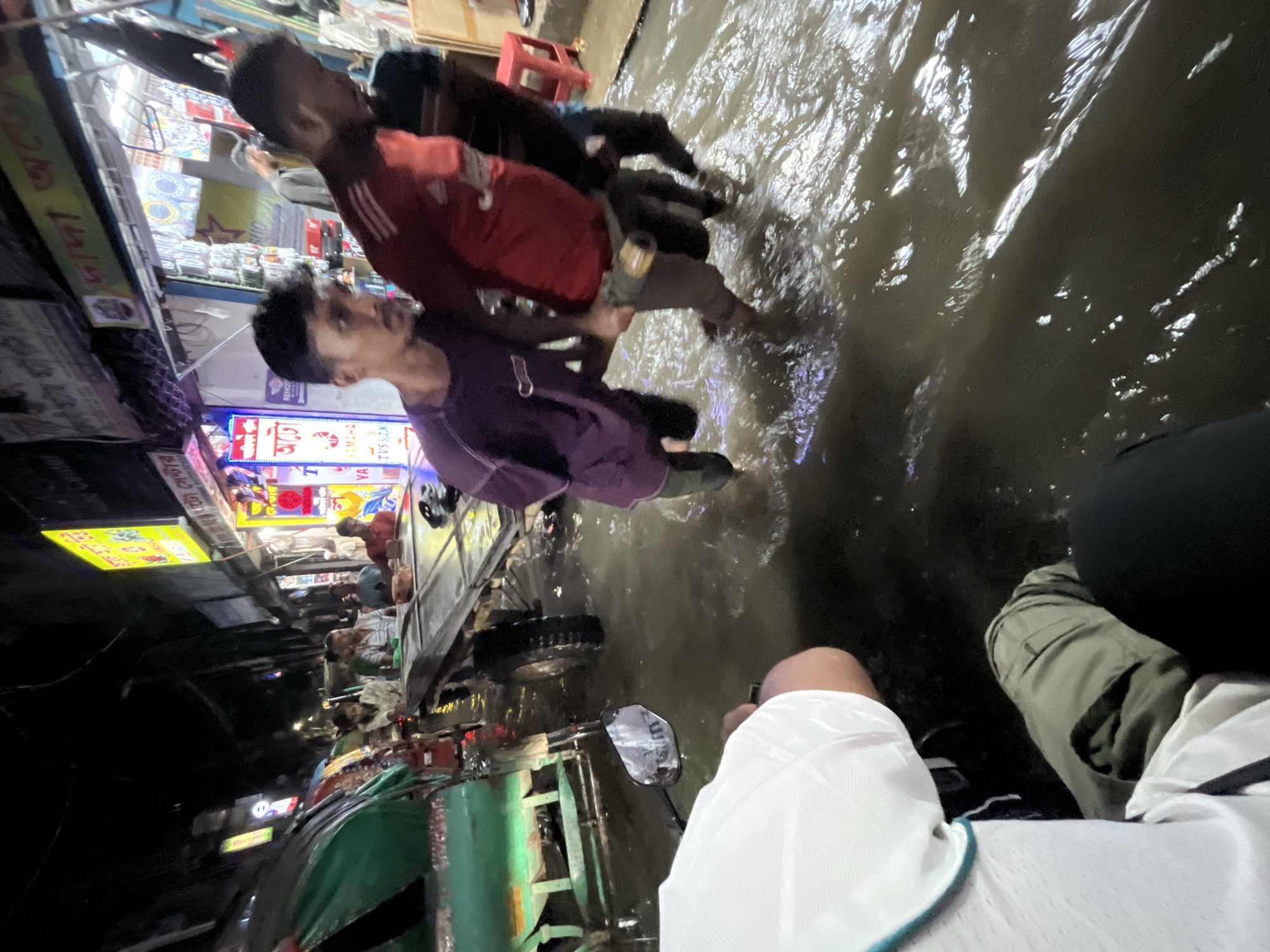 The image shows a flooded street scene in Dhaka, Bangladesh, likely during or after a period of heavy rain. The water level is high enough to submerge sidewalks and cover portions of the road. At least two men are wading through the floodwaters, one in a red shirt and the other in a purple shirt. The men are carrying objects, possibly helping others navigate the water. A rickshaw is partially visible, also in the floodwaters. The perspective is likely from the front seat of a vehicle. Storefronts with illuminated signs are visible along the street, indicating a commercial area.