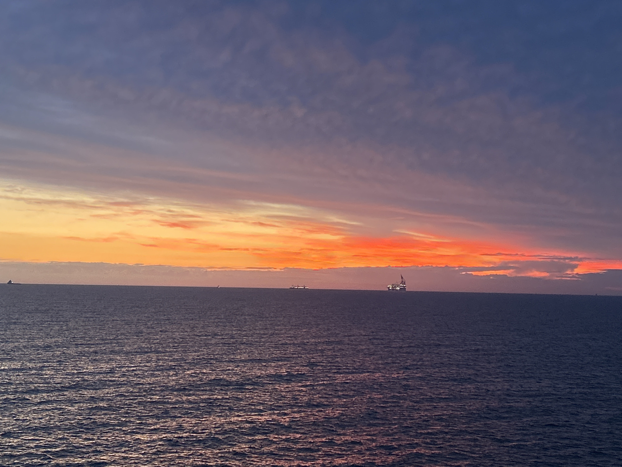 The image displays a panoramic view of a maritime sunset or sunrise over a large body of water in Unknown, Unknown. The sky exhibits a gradient of colors, transitioning from a deep blue-gray at the top, through shades of purple and soft pink, to intense bands of orange and fiery red along the horizon. Scattered, wispy clouds are present across the sky, particularly in the upper and middle sections, absorbing and reflecting the ambient light. The horizon line is straight and sharply defined, separating the dramatic sky from the calm ocean. The water surface is a dark, muted blue-gray, characterized by numerous small ripples and waves that catch subtle reflections. Several objects are visible on the horizon. On the far left, a dark silhouette of a large vessel is discernible. Towards the center-left, two additional, smaller ship silhouettes appear further in the distance. Prominently positioned on the right side of the frame is a large offshore platform or oil rig, illuminated with visible lights. A faint, small object may be present far to the right. No individuals are depicted in the scene.