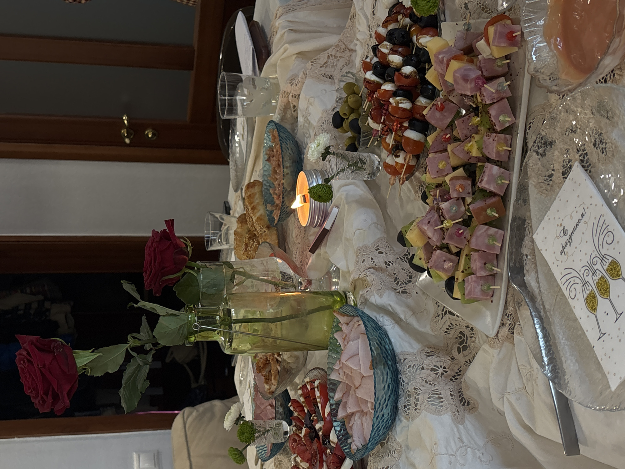 The image shows a buffet table laden with food, flowers, and decorations. There are several plates of appetizers, including skewers with cheese and ham, olives and cherry tomatoes, and sliced meats in decorative bowls. A candle illuminates the scene, and a vase of red roses is prominently displayed. A card with champagne glasses is also visible. The style of the decorations and food suggest a celebratory gathering. The photo was likely taken indoors. Information about the location in Krasnoyarsk, Russia, is not discernible from the image.