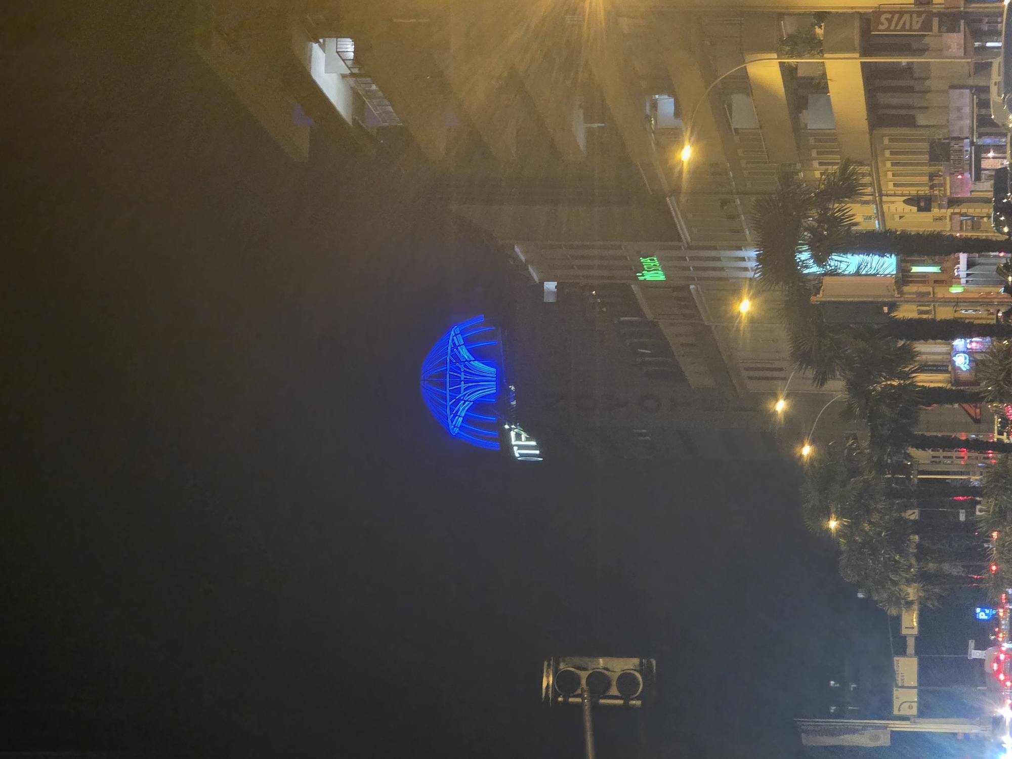 This is a night-time, low-angle, vertical photograph of an urban street scene in Montrouge, France, dominated by the illuminated dome of a building at the center, glowing bright blue. A traffic light is visible on the left side of the frame. Adjacent buildings line the street, with multiple floors and balconies, many windows are illuminated, casting a warm, yellow glow. The building with the blue dome has white lettering that appears to read "ITP". Below this, on another building, a green neon sign reads "ibis styles". A tall street lamp with a bright yellow light is prominent on the right, its light rays fanning out across the image. Several palm trees are sparsely planted along the sidewalk, with their silhouettes visible against the illuminated buildings. Traffic is visible in the foreground, with red taillights suggesting a slow or stopped flow of vehicles. The overall atmosphere is that of a city street at night, with artificial lighting creating a somewhat hazy or foggy effect. An "AVIS" sign is visible on a building towards the right.