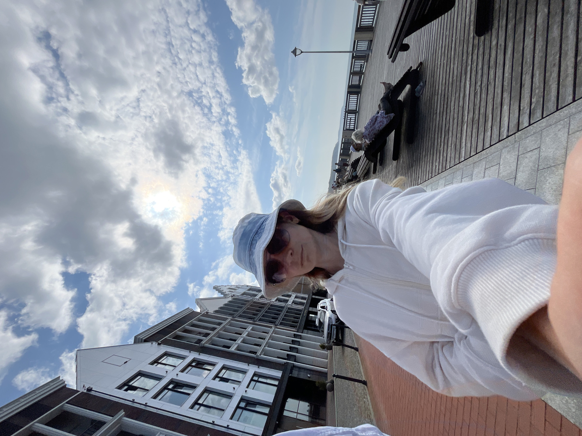 A young woman wearing a white hoodie and a light blue bucket hat is taking a selfie on a brick pathway. She is looking at the camera and her right arm is extended, holding what appears to be a phone. In the background, there are modern buildings with large windows, a white SUV parked, and a wooden boardwalk along the sea. Several people are visible on the boardwalk, some sitting on benches and others walking. The sky is partly cloudy with the sun visible. The scene is located in Svetlogorsk, Russia, as indicated by the presence of a street lamp and a white SUV with Russian license plates.