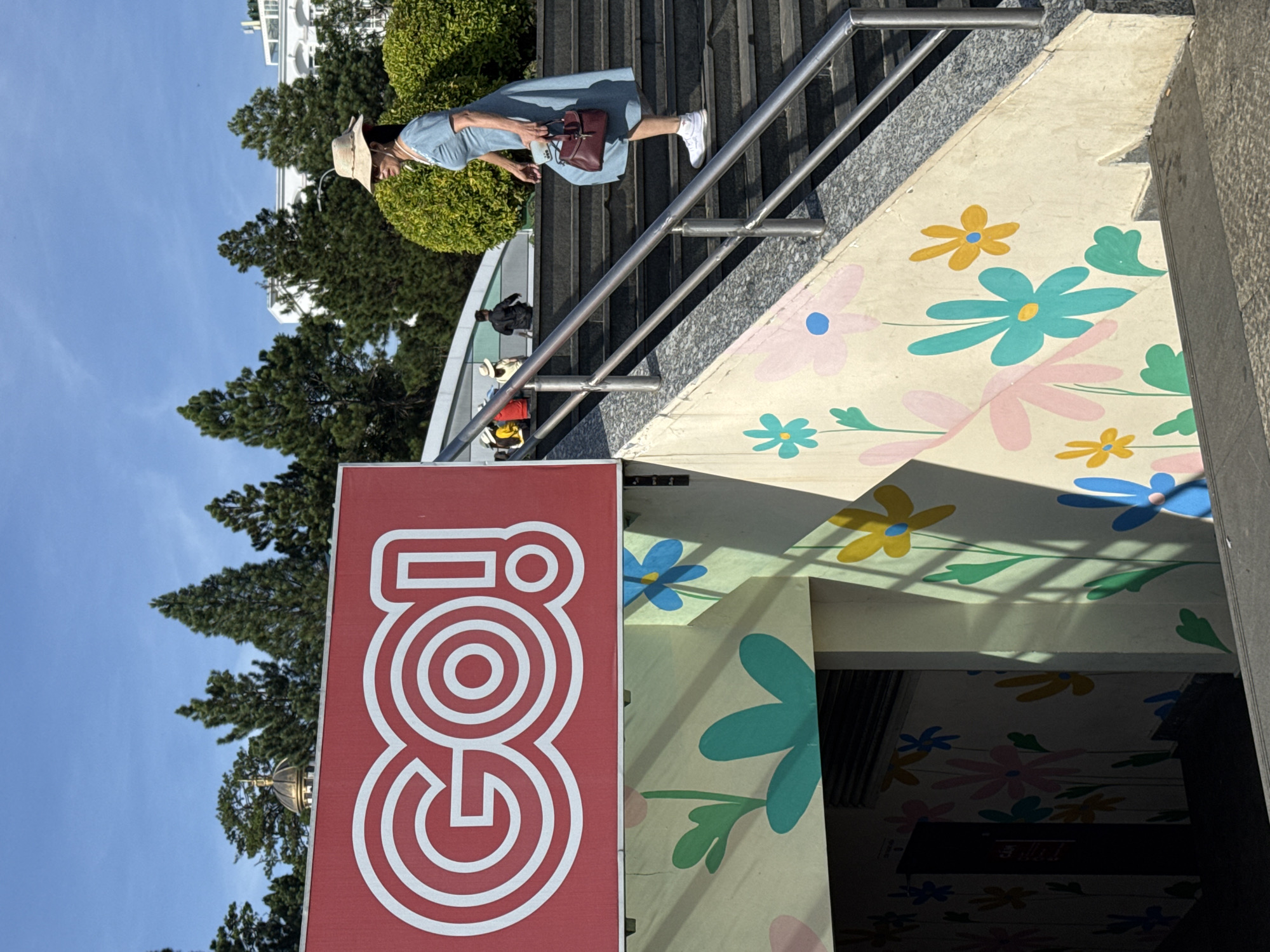 In Da Lat, Vietnam, a woman in a blue dress and a straw hat ascends a concrete staircase beside a vibrant mural of large, stylized flowers. She carries a small, dark red handbag and wears white sneakers. Above the stairs, a red sign with white lettering displays "GO!". The background features manicured green bushes, tall evergreen trees, and distant white buildings under a clear blue sky. The sun casts distinct shadows across the floral artwork on the wall.