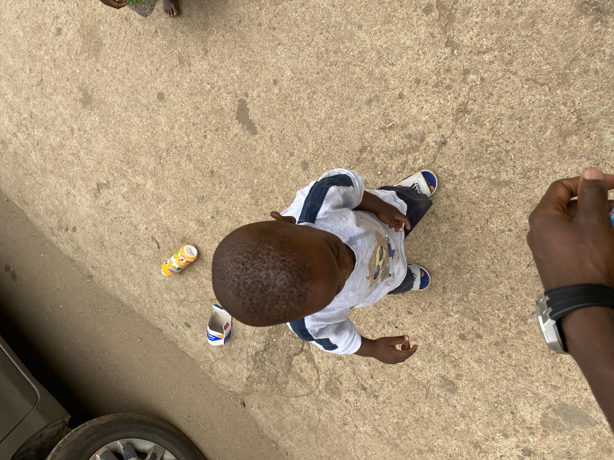 A top-down shot shows a young child standing on a concrete surface in Bukuru, Nigeria. The child is wearing a white shirt with blue sleeves and dark shorts, along with blue and white sandals. To the child's left is an empty carton of "Choco Malt" and a partially eaten can of "Bobo" juice. A portion of a car tire is visible in the lower-left corner of the frame. An adult arm with a wristwatch is visible on the right side of the frame. The background is a textured concrete surface. Another foot is visible in the top of the frame.