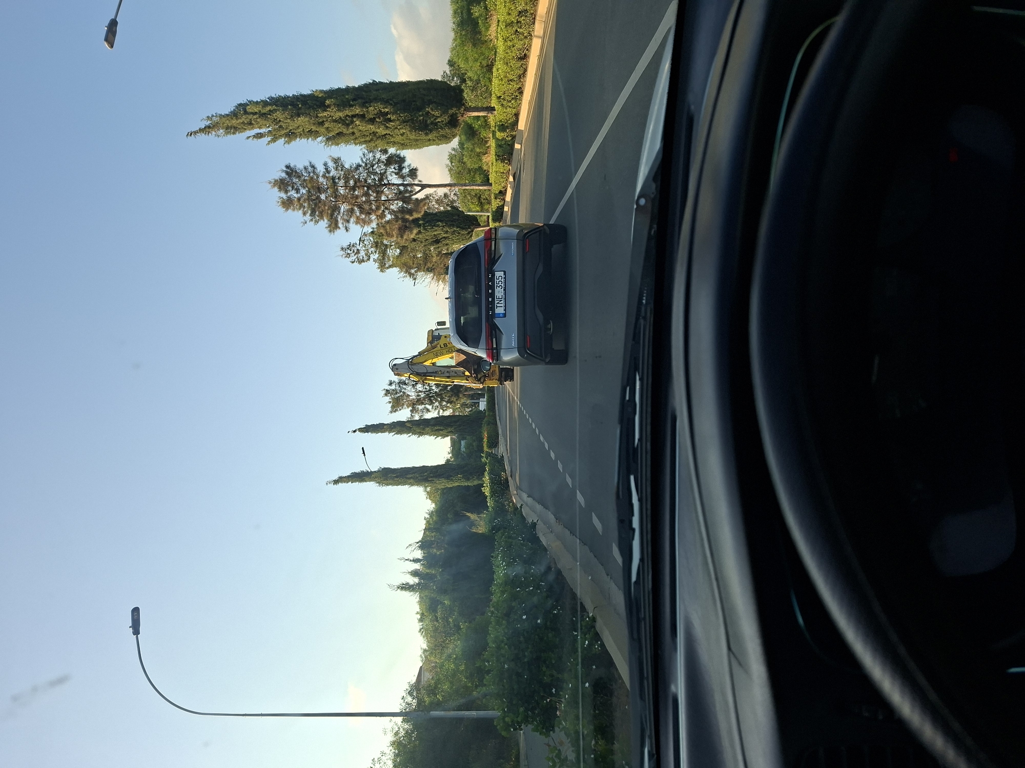 The image, taken from inside a vehicle, shows a road in Protaras, Cyprus. A silver Nissan vehicle with license plate TNE 355 is visible in front. Further ahead, a yellow construction excavator is parked. Trees and street lights line the road. The sky is blue.