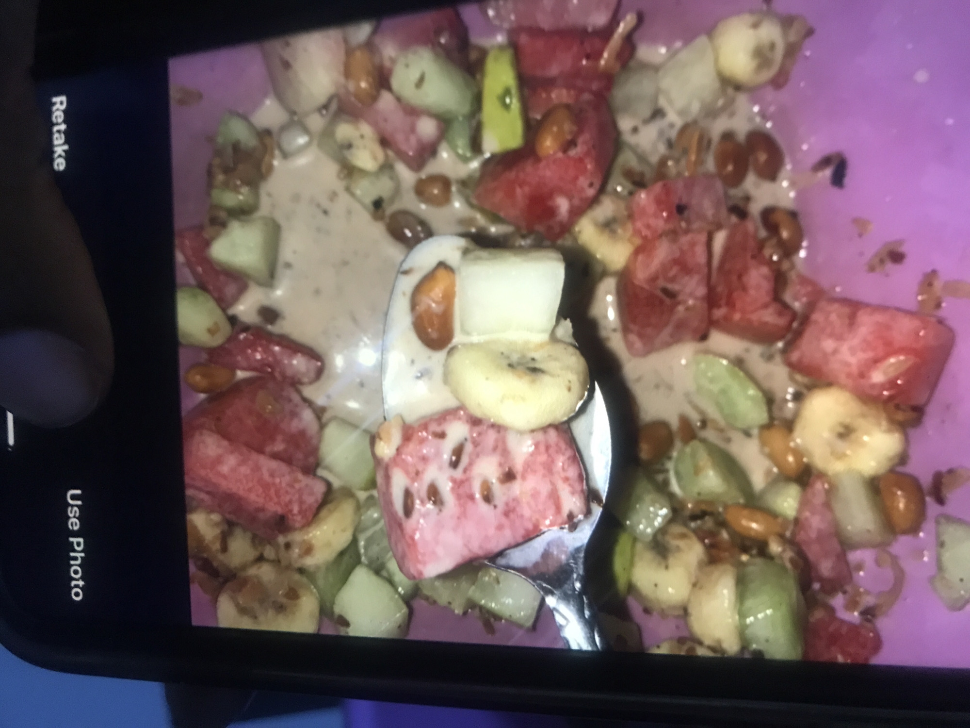 The image displays a close-up shot of a smartphone screen held horizontally by a person's left hand, with a thumb resting on the side. The phone's camera interface is visible, showing a freshly captured photo of a food dish. Vertically oriented text "Retake" is visible on the left side of the screen, and "Use Photo" on the bottom right, indicating options for the recently taken picture.
The main subject of the photo on the screen is a vibrant bowl of food, likely a fruit salad or smoothie bowl, presented in a purple container. The dish contains a mixture of chopped fruits, nuts, and a creamy, light brown liquid base. Recognizable fruits include chunks of red fruit (possibly watermelon or strawberry), slices of banana, and pieces of light green fruit (resembling cucumber or kiwi). Small, brownish nuts or seeds are scattered throughout the mixture. A silver spoon is positioned in the center, scooping up a portion of the food, which features a piece of the red fruit, banana slices, and some nuts. The overall appearance suggests a chilled or frozen preparation.
The activity depicted is the act of photographing food with a smartphone. The background surrounding the phone is dark and out of focus, offering no specific details about the immediate surroundings beyond the phone and hand. The scene takes place in Rafin Par, Nigeria.