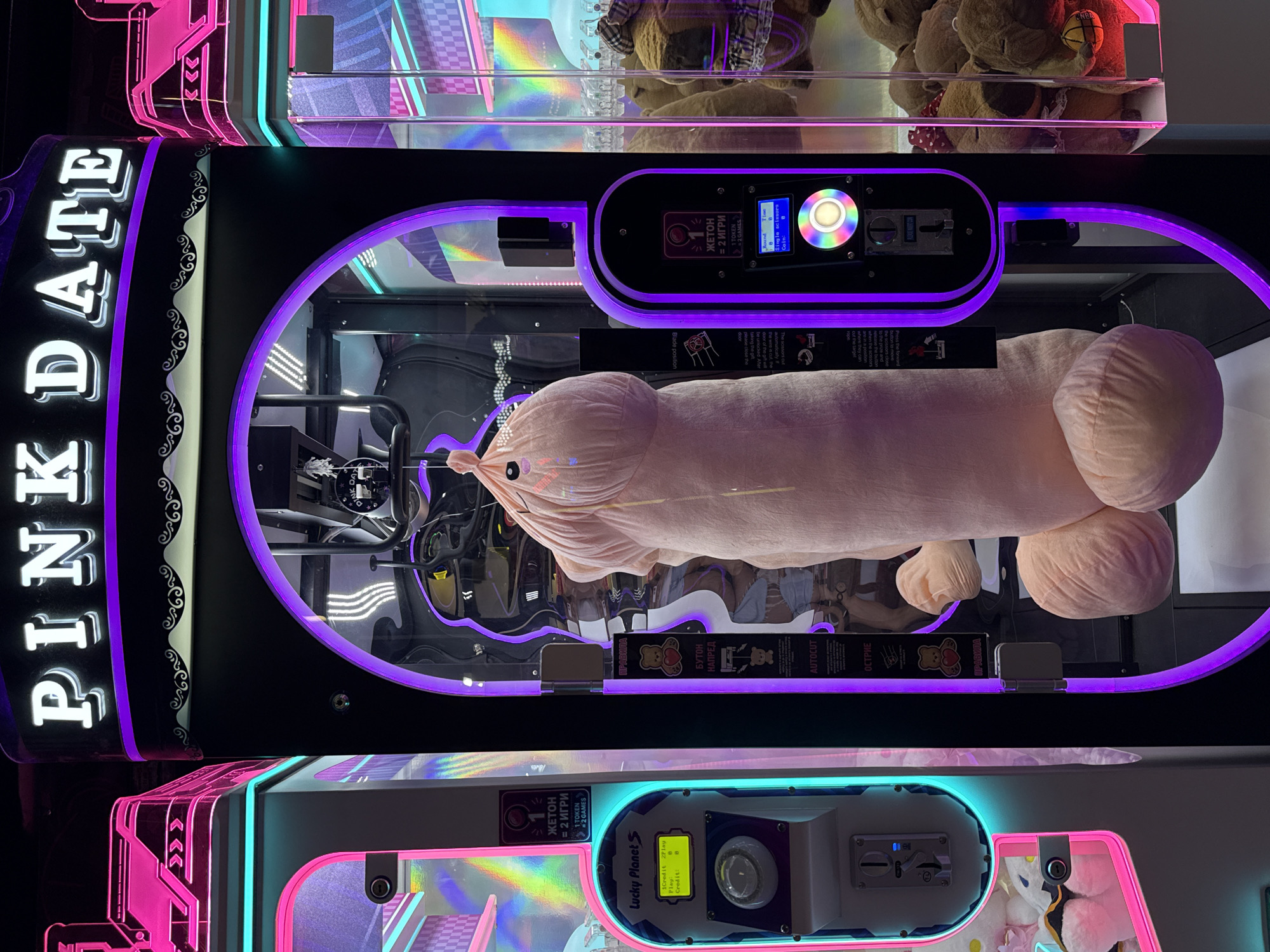 This image shows a close-up of a "PINK DATE" claw machine arcade game, featuring a large, plush phallic-shaped toy as the prize. The machine is illuminated by neon pink and purple lights. On the left side of the machine, there's a "Lucky Planet" display showing game credit and play information. On the right side, there's another coin slot and display, with instructions in Russian. The background of the machine's interior shows a rainbow-colored checkered pattern and more plush toys. The overall environment appears to be an indoor arcade or entertainment venue. The image is taken from a frontal perspective, with no people visible in the frame.