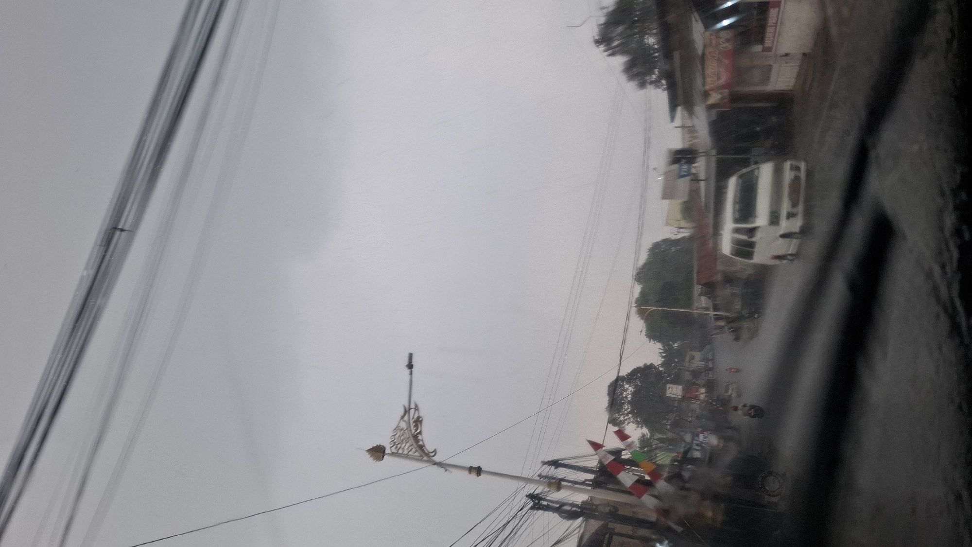 The image depicts a rainy street scene in Cilaku, Indonesia. A white van is driving down the wet road, with rain visibly falling. To the left, a decorative street lamp stands tall, adorned with intricate golden scrollwork. Below it, a series of flags in red and white are strung across, suggesting a local event or celebration. Buildings line the street, some with visible signage. The sky is overcast and a muted gray. The perspective is from inside a vehicle, with raindrops distorting the view through the windshield.
