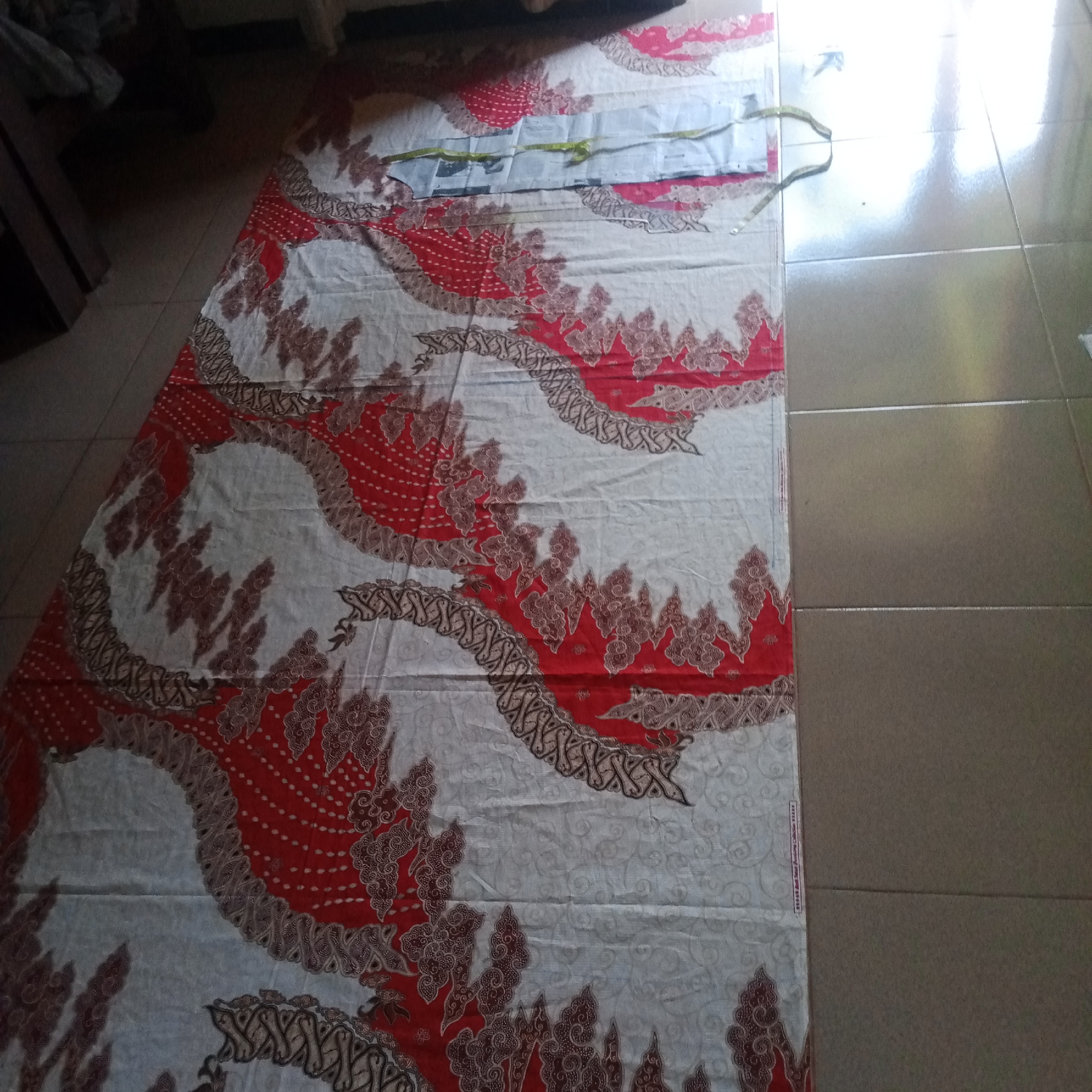 In the village of Semanding, Indonesia, a long piece of batik fabric is spread out on a tiled floor. The fabric features a striking pattern of red and white with intricate brown detailing. The pattern is dominated by large, circular motifs in red, filled with small white dots and surrounded by elaborate brown lace-like borders. Interspersed between these are flame-like shapes in brown against the white background, also outlined with the detailed brown border. The fabric appears to be laid out for cutting or display, with a segment of lighter fabric and a green measuring tape visible near the top. The tiled floor, likely a common surface in Indonesian homes, consists of large, neutral-colored tiles that reflect the ambient light, indicating an indoor or covered outdoor setting. The overall scene suggests a domestic or artisanal environment where fabric is being worked with.