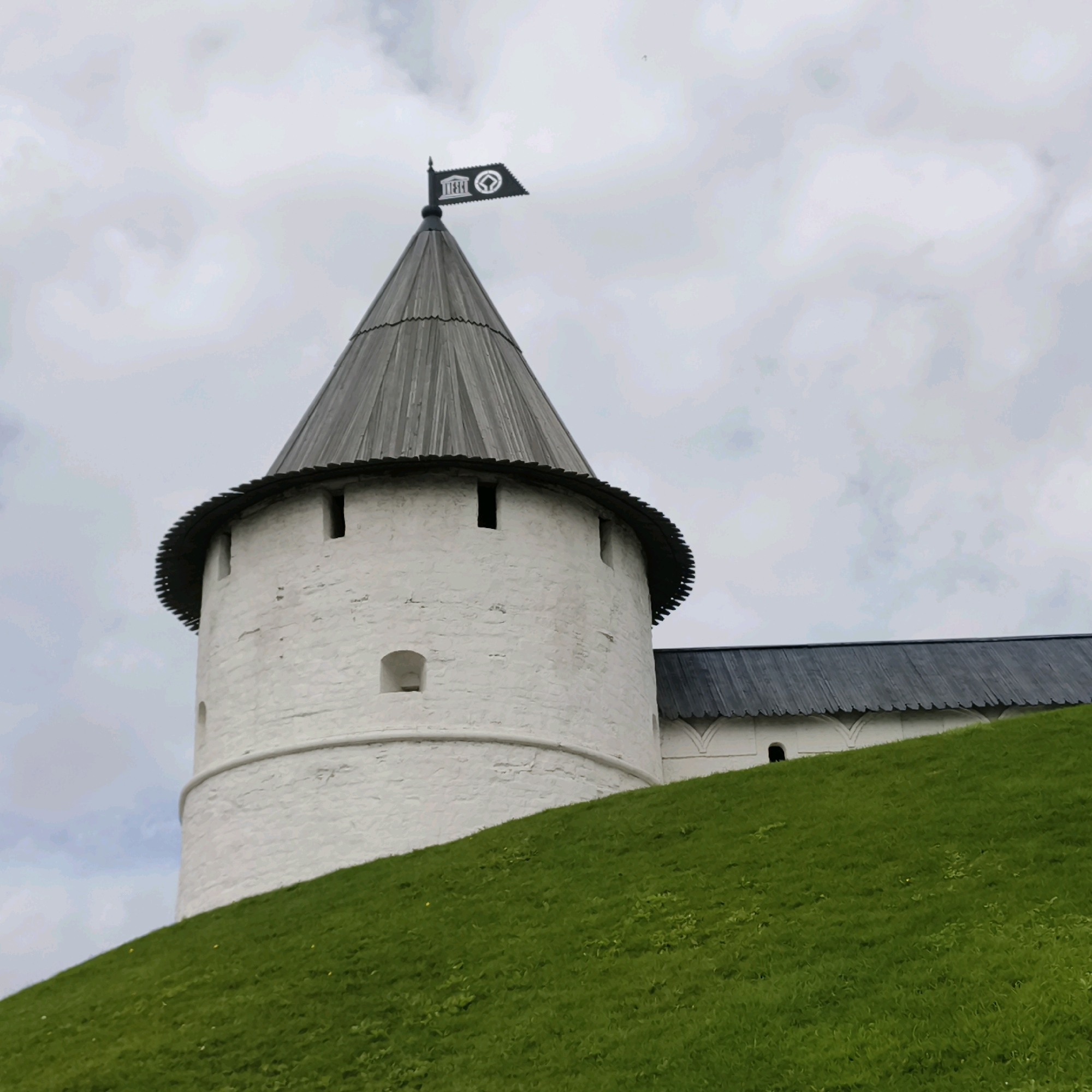 The image shows a view of the Kul Sharif Mosque in Kazan, Russia, on a cloudy day. A white brick tower with a conical wooden roof stands in the foreground, with a black flag on top. A grassy hill slopes up towards the tower. The Kul Sharif Mosque is a prominent landmark in Kazan, and this view captures its architectural beauty. The mosque is located in the Kazan Kremlin, a UNESCO World Heritage Site.