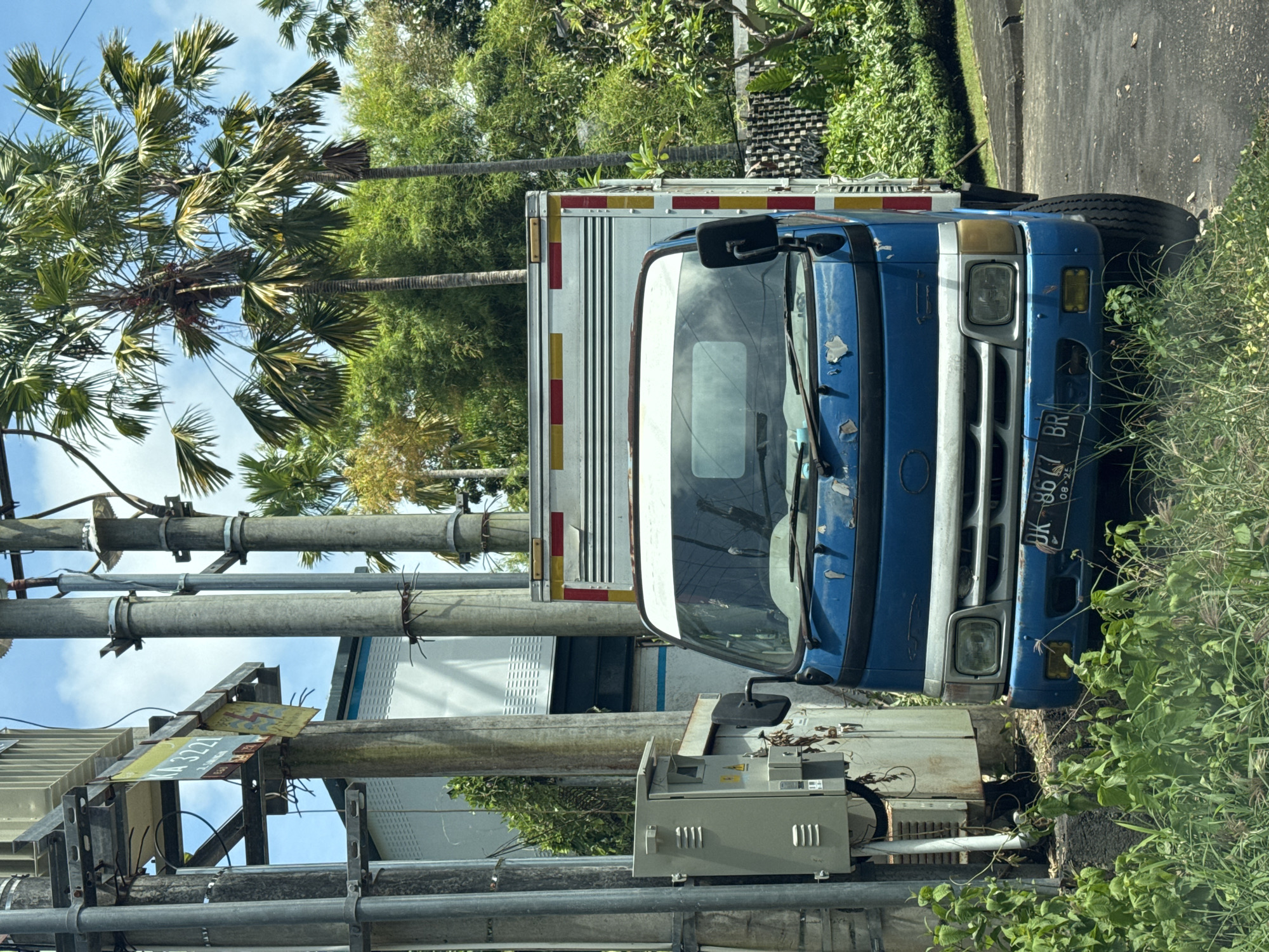A blue truck with a white cargo box is parked along a road in Nusa Dua, Indonesia. The truck has a license plate that reads "DK 8617 BR." A utility pole with electrical equipment is located next to the truck. Palm trees and other greenery are visible in the background. The road is paved and appears to be in a residential or commercial area.