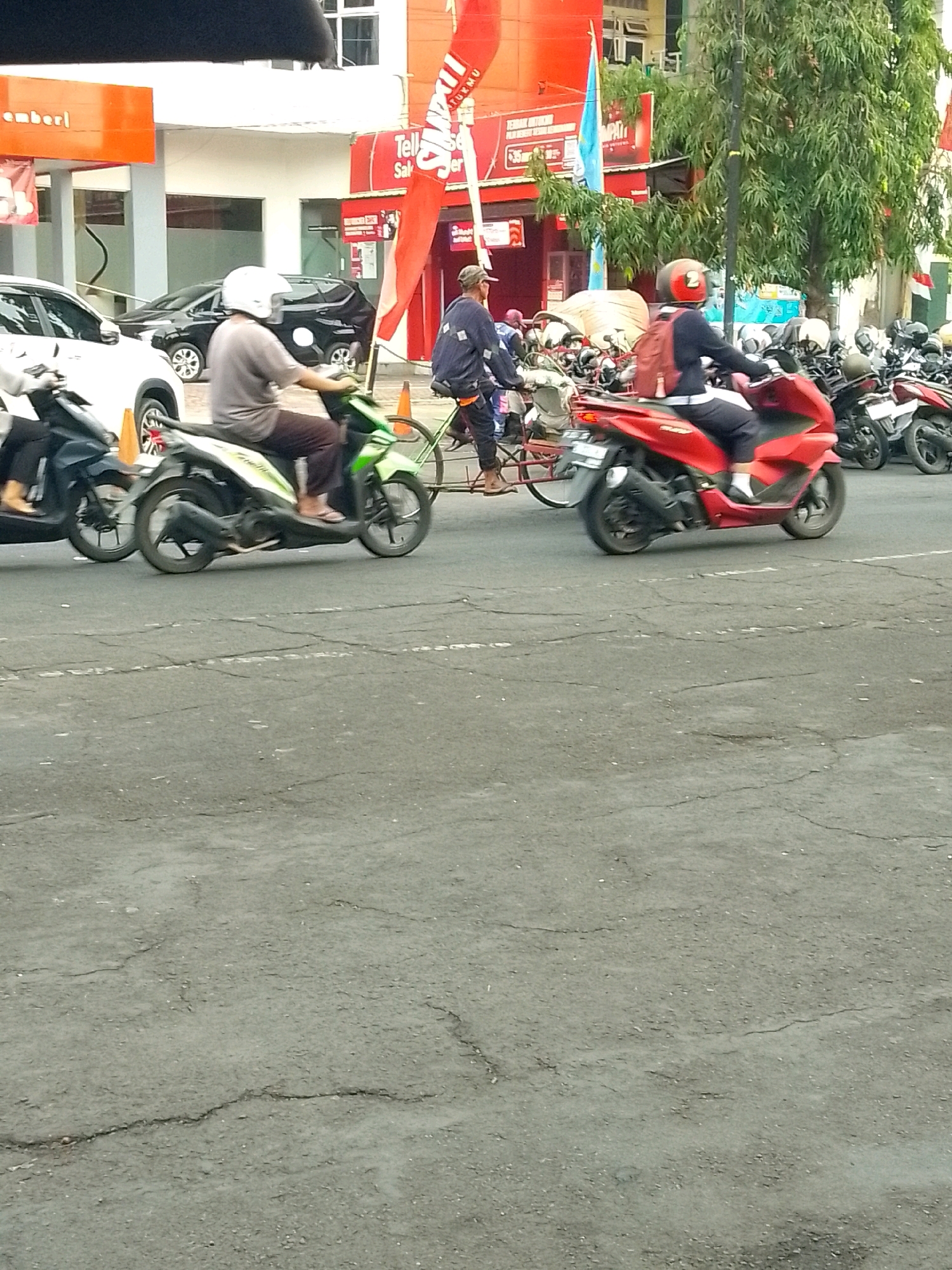 The image shows a street scene in Jember, Indonesia, likely on a busy day. In the foreground, there is a cracked asphalt road. Several motorbikes are visible, including a green one with a rider in a gray shirt and white helmet in the left middle ground, and a red scooter with a rider in a dark jacket and red helmet in the right middle ground. Behind these motorbikes, a person is riding a bicycle. The background features buildings with orange and red signage, including what appears to be a business or store with the word "emberl" prominently displayed. Trees with green foliage are also present. Several parked motorbikes are visible in the distance. The overall impression is of a typical urban environment with active traffic.