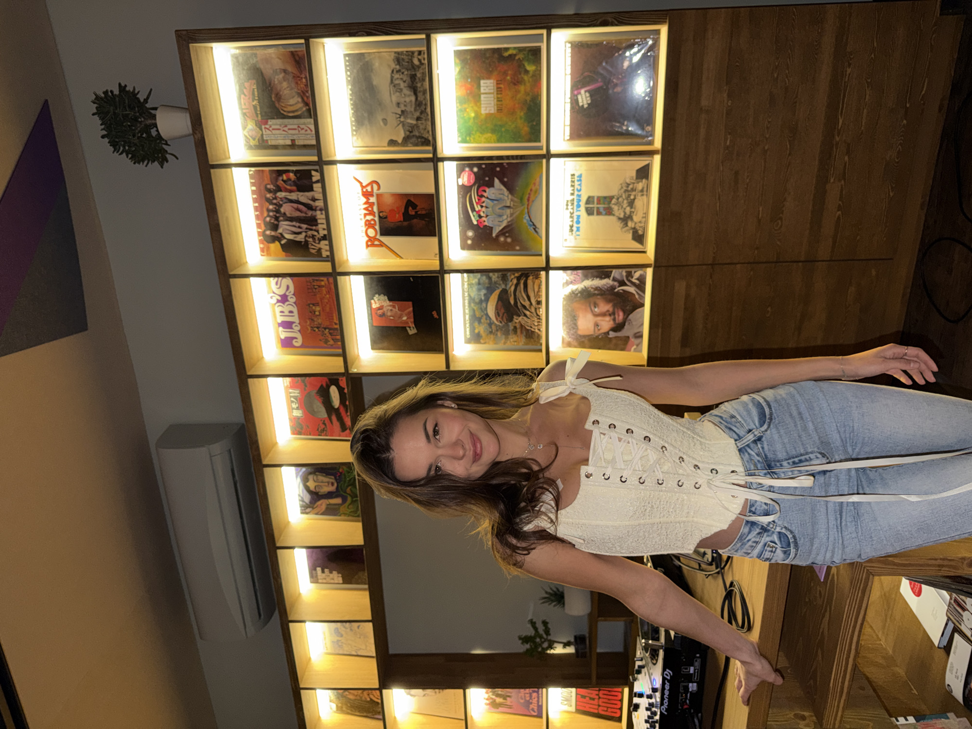 An indoor portrait photograph captures a young woman smiling cheerfully at the viewer, positioned in what appears to be a music studio or a dedicated listening space. She has long brown hair and is dressed in a white, corset-style lace-up top paired with light blue jeans.
Behind her, a prominent wooden shelving unit, brightly illuminated by warm LED strips, showcases a collection of vinyl records displayed face-out. Recognizable album covers include "SUGARCANE HARRIS," "BOBIAMES" (Bob James), "I'M ON YOUR CASE" (The Meters), and "J.B.S." (likely James Brown's Funky People). Other visible text on record sleeves includes "初モノ" (Hatsu Mono) and fragments like "Chics," "SMOO," "HEA," and "Cla." A small potted plant rests on the top shelf. Adjacent to the woman, a Pioneer DJ mixer, bearing "Pioneer DJ" and "GOOD" branding, sits on a wooden counter, indicating a setup for music production or DJing.
The room's left wall features an air conditioning unit and a purple and grey geometric artwork. The warm lighting suggests an evening or nighttime setting. The woman's pleasant expression creates an inviting and relaxed ambiance within this music-centric environment, located in Krasnoyarsk, Russia.