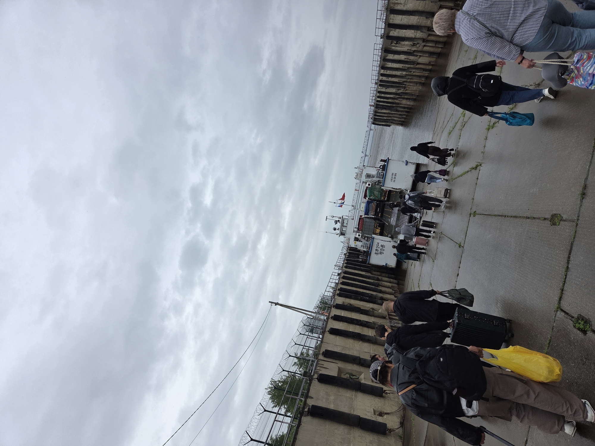 The image captures a bustling scene on a concrete dock under a heavily overcast sky, likely in Nizhneleninskoe, Russia. Numerous individuals, mostly viewed from behind, are seen walking along the wet-looking ground, laden with luggage including backpacks, wheeled suitcases, and various bags. Their attire, consisting of jackets and long pants, suggests cool or damp weather.
The primary activity appears to be people preparing to board a white-hulled boat, possibly a ferry or river vessel, which is moored alongside the dock. The boat displays visible text on its side: "DRAGOR 004", "КН ЕР ВЫН", and "МНД". A sign on the boat also reads "ЧИСТОЕ ПОМЕЩЕНИЕ" (Clean Room). Red and white flags are visible on the boat's mast.
The concrete dock features prominent black rubber fenders along its edge, intended for ship berthing. A metal fence topped with barbed wire runs along a section of the dock, separating the pedestrian area from what might be an adjacent restricted zone or industrial facility. In the distance, across the wide body of water, a faint bridge structure is discernible. The cloudy sky, characteristic of an overcast day, fills a large portion of the upper frame, contributing to the muted lighting of the scene. The overall impression is one of purposeful movement and transit, with individuals focused on their journey.