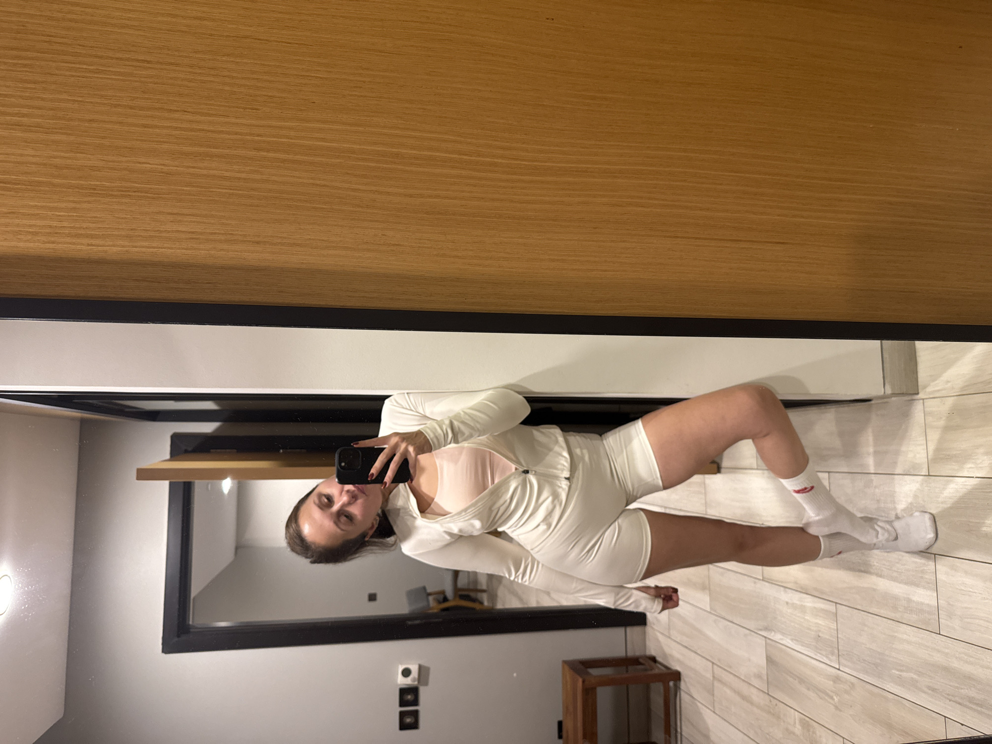 A woman is captured in a mirror selfie, standing indoors and holding a black smartphone to take the picture. She is wearing a light-colored (likely white or off-white) zip-up jacket over a light pink or beige top, and matching light-colored shorts. On her feet, she wears white socks with a red Nike swoosh logo visible on her right sock.
The large mirror, framed in black, reflects the woman and the room around her. In the reflection, the floor is covered with light-colored rectangular tiles arranged in a staggered pattern. A small wooden table or stool with slender legs is visible in the lower right corner of the reflection. On the wall behind her in the reflection, there are two dark rectangular elements, possibly outlets or light switches, and a round wall-mounted light fixture further to the left. Above the mirror, a light brown wooden panel or shelf extends horizontally. The setting appears to be a modern indoor space, potentially a hotel room or apartment in Dubai, United Arab Emirates, illuminated by artificial light.