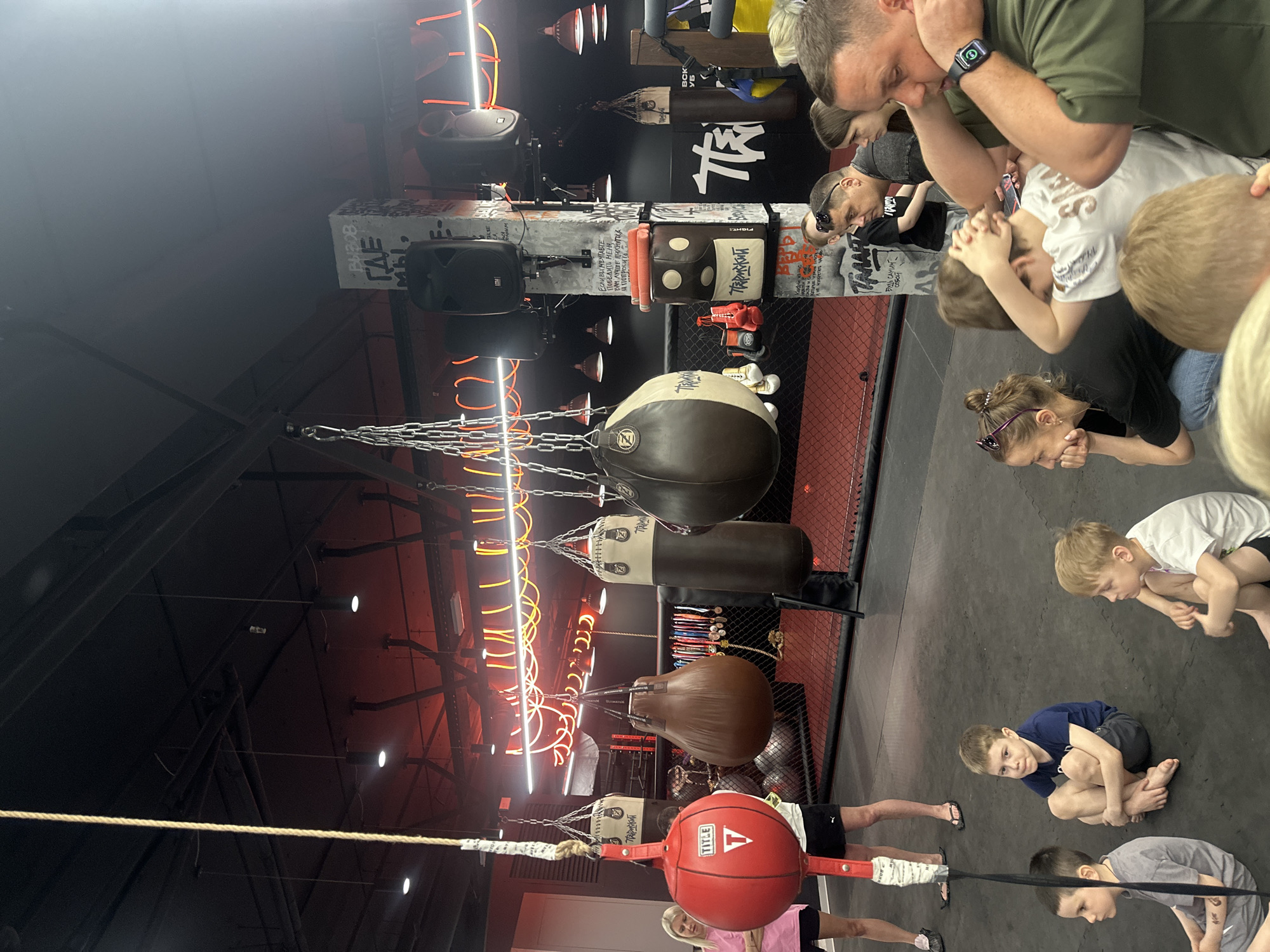 A group of children and adults are in a boxing gym, possibly in Perm, Russia, given the Cyrillic text visible on the equipment. Various punching bags, including a red "TITLE" speed bag, are hanging from the ceiling. Neon lighting is present. People appear to be observing or participating in a boxing demonstration.