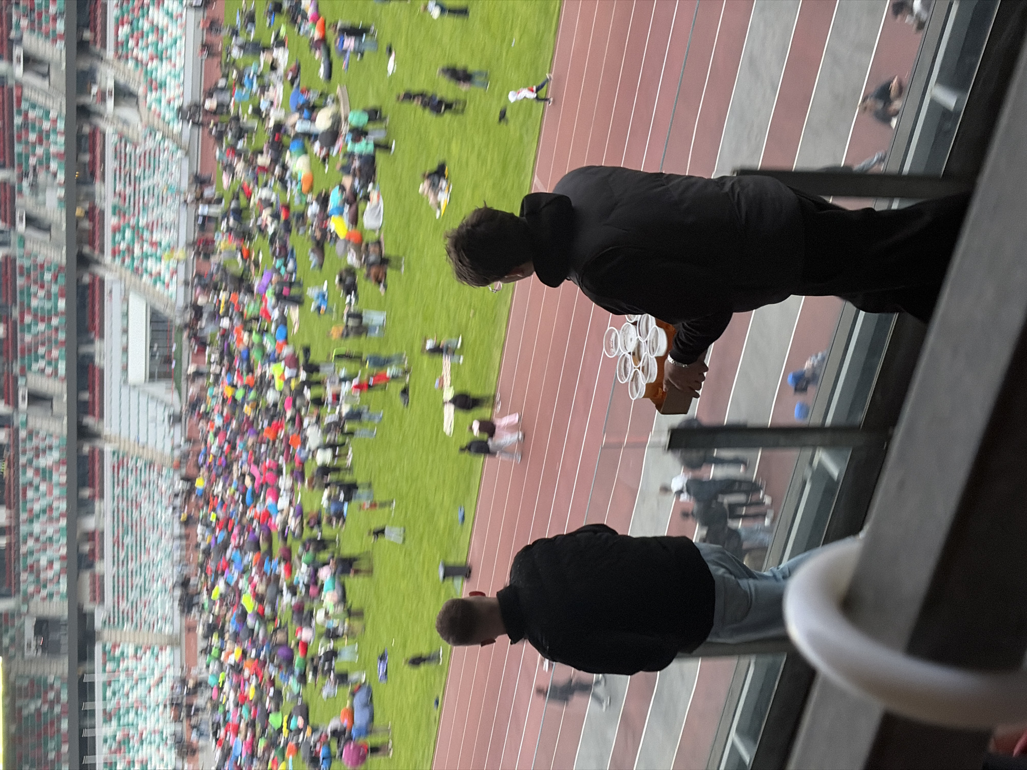 From an elevated vantage point, the image depicts a stadium scene in Minsk, Belarus. The stadium features a large crowd gathered on a central green space, surrounded by running tracks. Spectator seating is visible in the background. Two individuals are positioned in the foreground, presumably on a balcony or within an enclosed area of the stadium. One individual is holding a stack of cups, possibly containing beverages. The architecture suggests the scene is indoors, as there are windows or glass barriers between the individuals and the stadium activity.