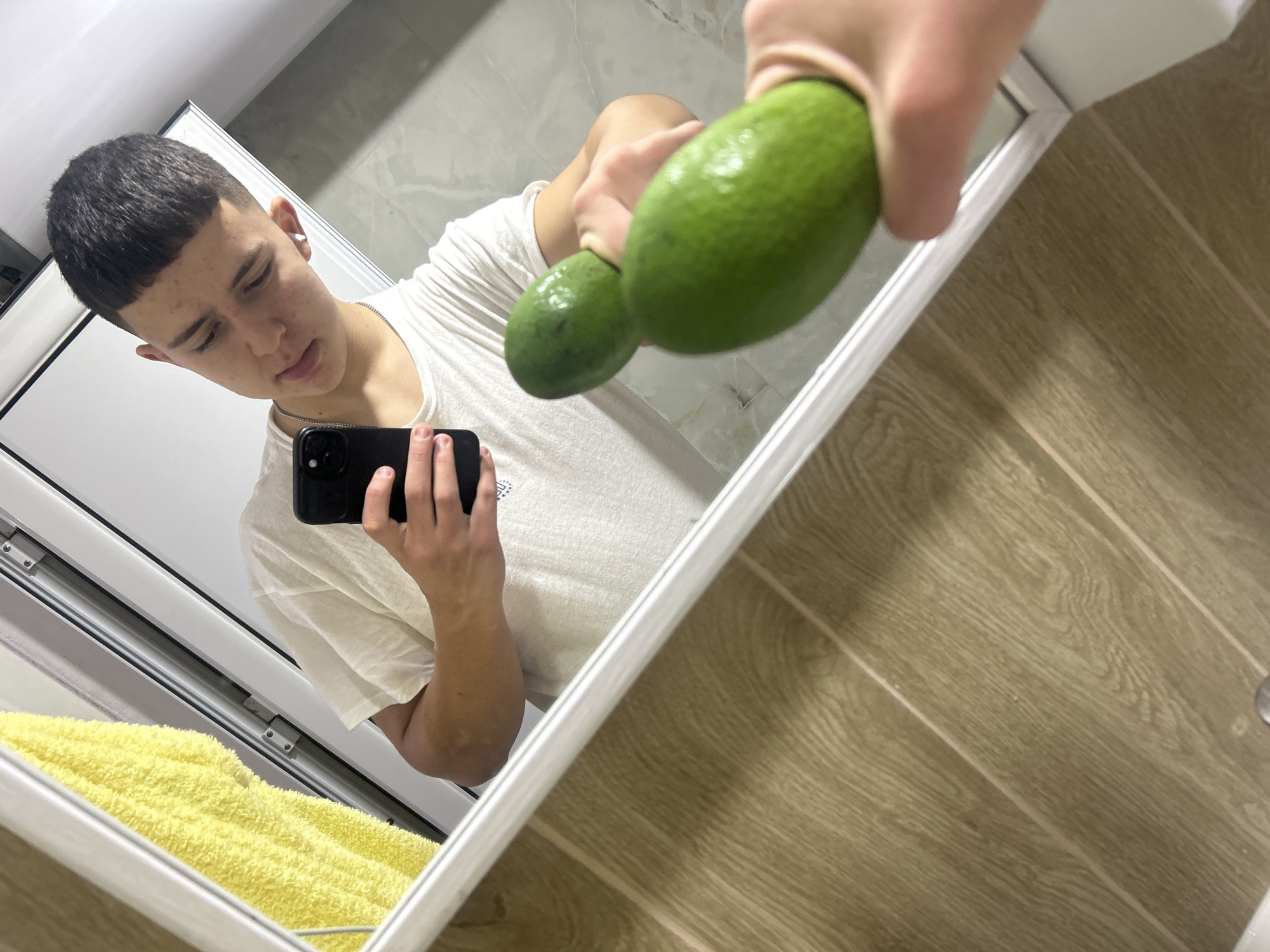 This image depicts an indoor scene in Zlatograd, Bulgaria, captured as a mirror selfie. A young male subject, with short dark hair and wearing a light-colored t-shirt and a silver chain, is visible in reflection. He is holding a black smartphone in his right hand (as reflected) and two elongated green avocados in his left hand (as reflected). The avocados appear moist. The reflected background features a yellow terry cloth towel hanging on the left, a white-framed door or enclosure, and a light-toned, subtly patterned tiled wall. The lower foreground shows a segment of a light brown, wood-patterned floor directly beneath the mirror frame.