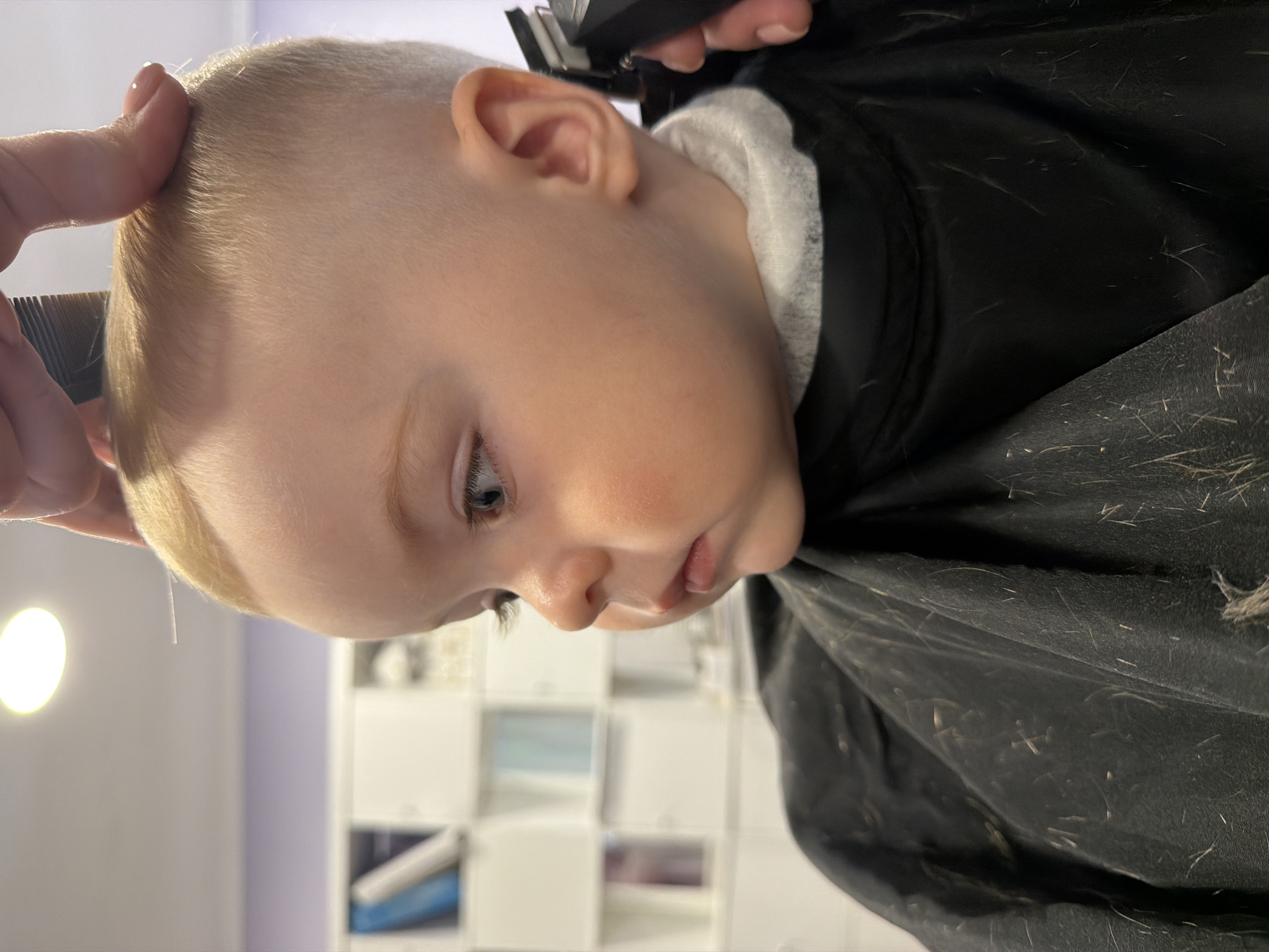 A young child is getting a haircut. The child has short blond hair, blue eyes, and fair skin. They are wearing a black cape over a white shirt. Hair clippings are visible on the cape. A person is holding a comb in one hand and a hair clipper in the other, cutting the child's hair. The background shows a white cabinet with items on its shelves. The location is unknown and cannot be determined to be Votkinsk, Russia based on the image.