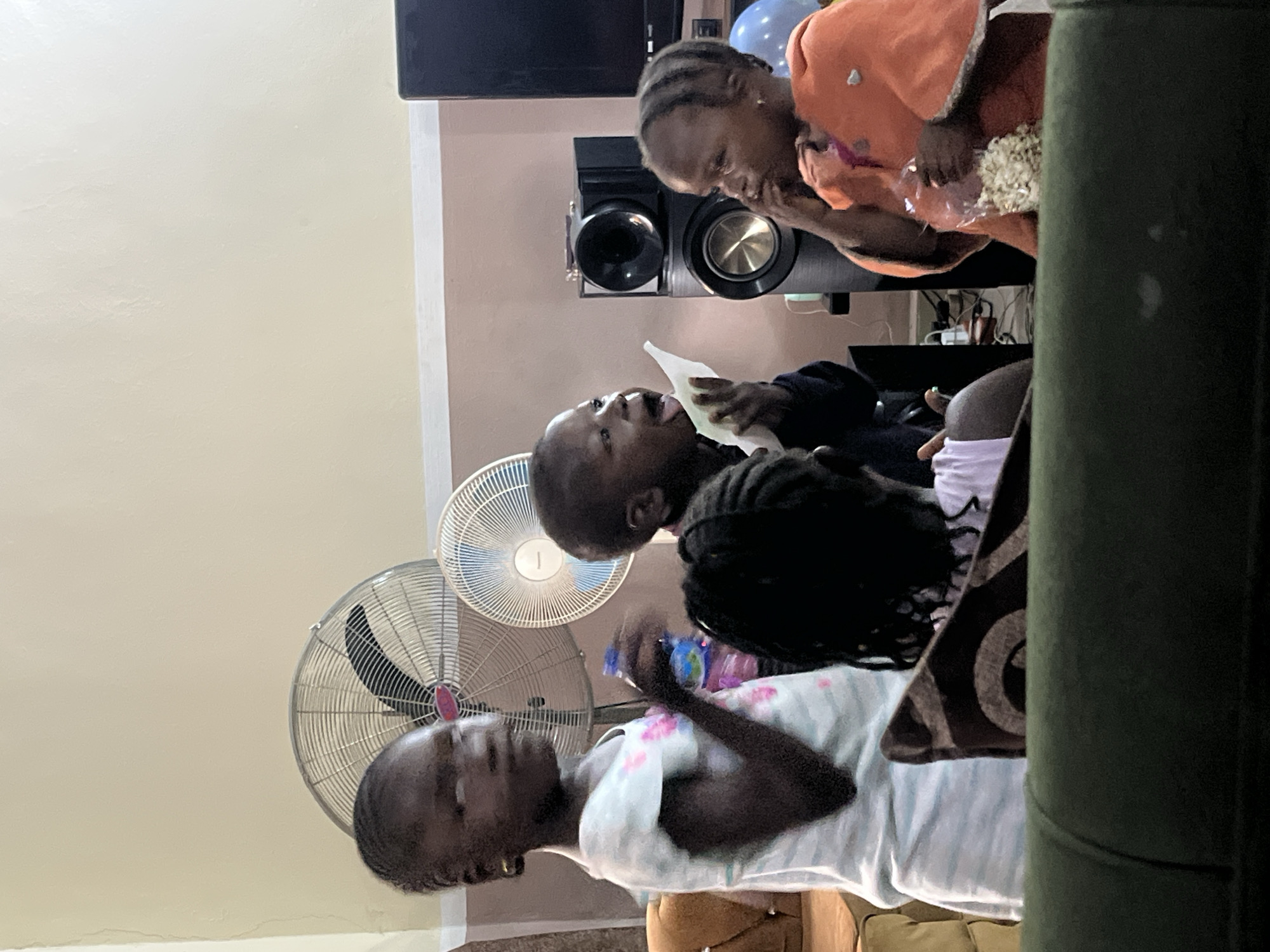 In Bukuru, Nigeria, a group of four young children are gathered in a room, possibly a living area. The children, appearing to be between the ages of three and eight, are positioned in the center of the frame. Two of the children are actively engaged with food items, with one child holding a bag of popcorn and another with an open mouth, possibly in the process of eating. There are two fans visible in the background, suggesting a warm climate. A television and speakers are also present, indicating a space equipped for entertainment. The children are dressed casually in t-shirts and a hooded garment. The lighting appears to be artificial, contributing to the indoor setting.