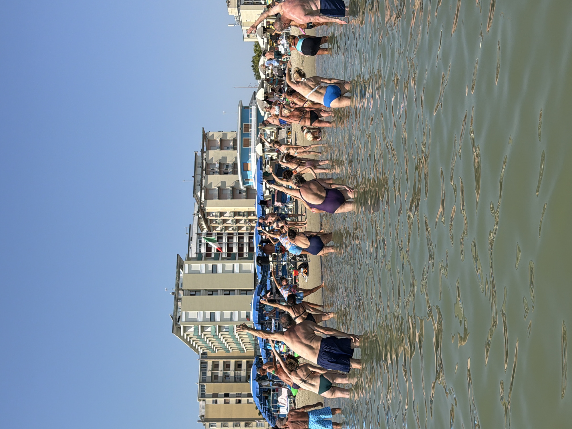 A group of people are doing exercises in the shallow water of the Adriatic Sea on the beach in Gatteo Mare, Italy. The people are positioned in a line, facing away from the camera, and are stretching their arms and torsos. They are wearing various types of swimwear. In the background, there are several buildings that appear to be hotels or apartment complexes, beach umbrellas, and lounge chairs. The sky is clear and blue.