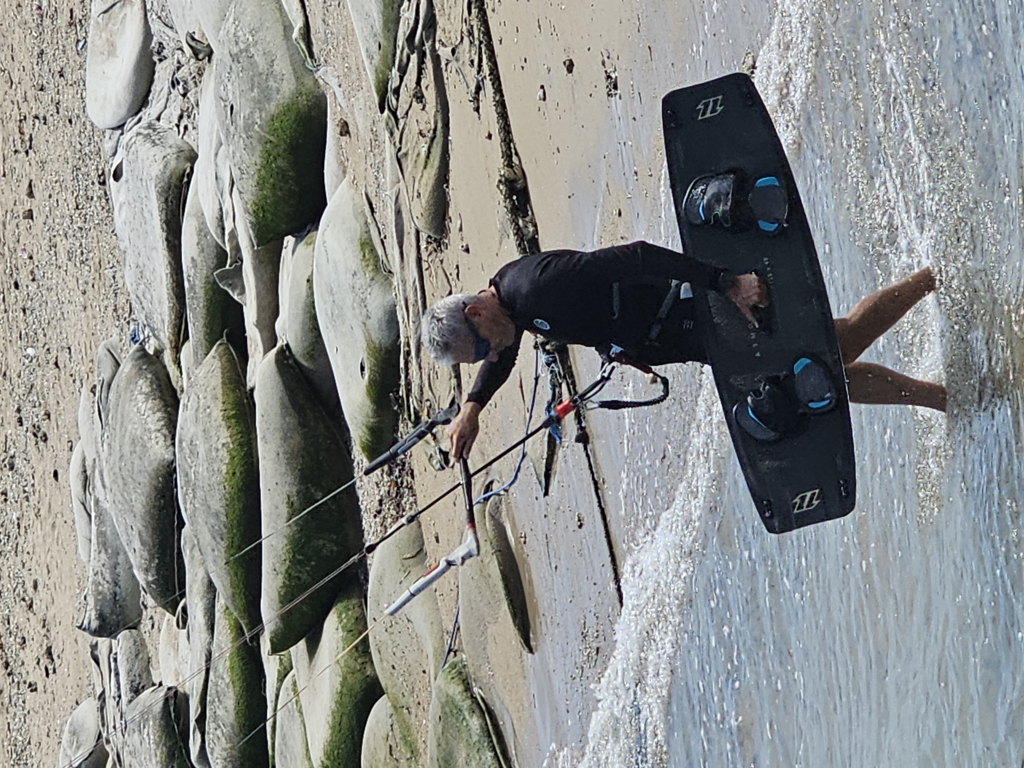 An older Caucasian male with graying hair and sunglasses is walking in shallow water near a beach. He is wearing a black wetsuit and carrying a black kiteboard. The kiteboard is oriented horizontally and has bindings attached. The man is holding a kite control bar in his right hand, and the kite lines are visible extending upwards. The beach is sandy and rocky, with large, moss-covered boulders scattered along the shoreline. The water is clear and has small waves lapping at the shore. The scene appears to be an outdoor recreational area.