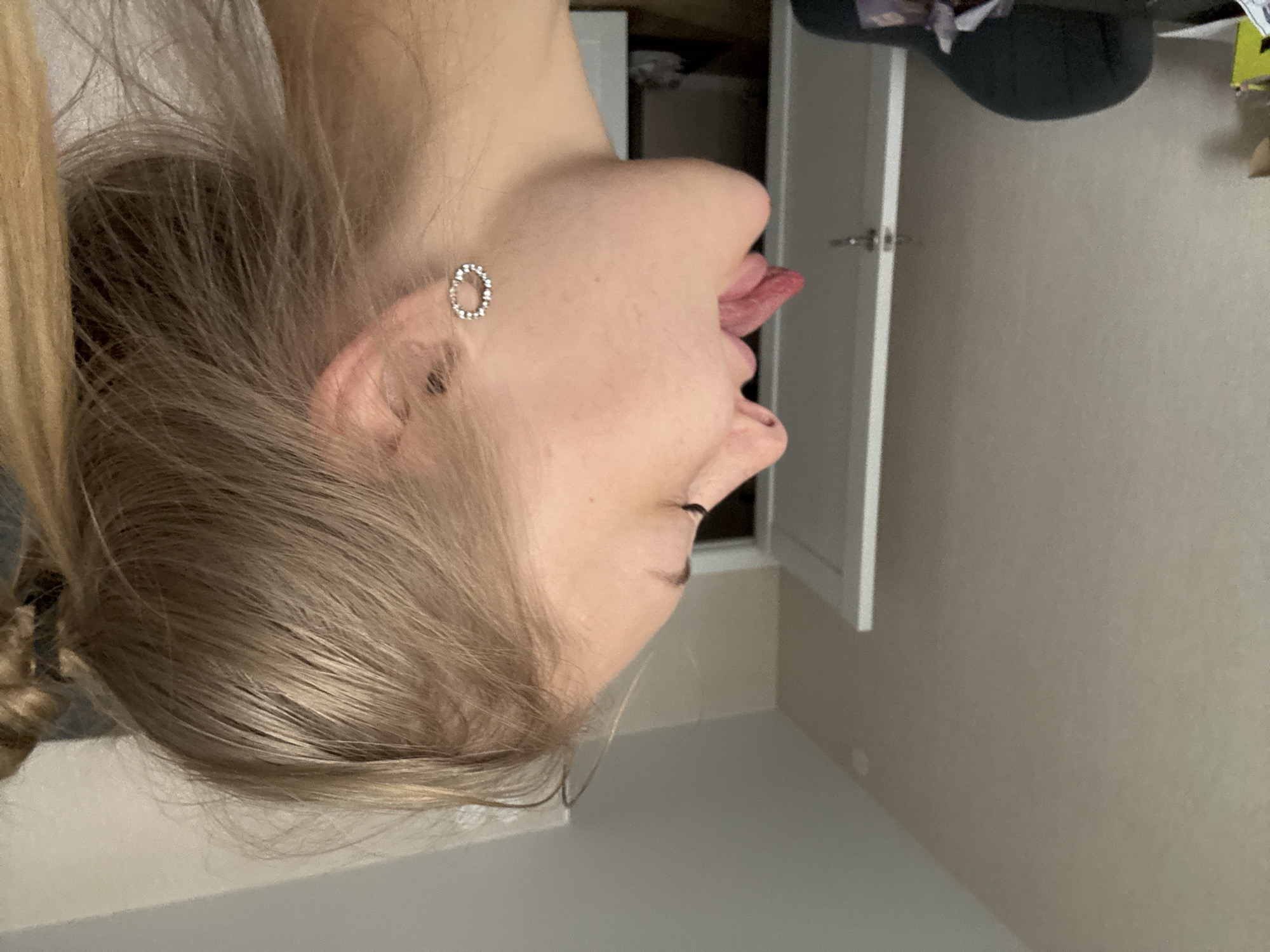In a dimly lit room, a young woman of Caucasian ethnicity, with blonde hair pulled back into a ponytail and a small circular earring adorned with rhinestones, is captured in profile. Her tongue is extended, and she appears to be making a playful gesture. The background reveals a neutral-colored wall, a white door with a silver handle, and a portion of a black chair. The scene is domestic, suggesting an indoor setting. There is no discernible connection to the city of Perm, Russia.