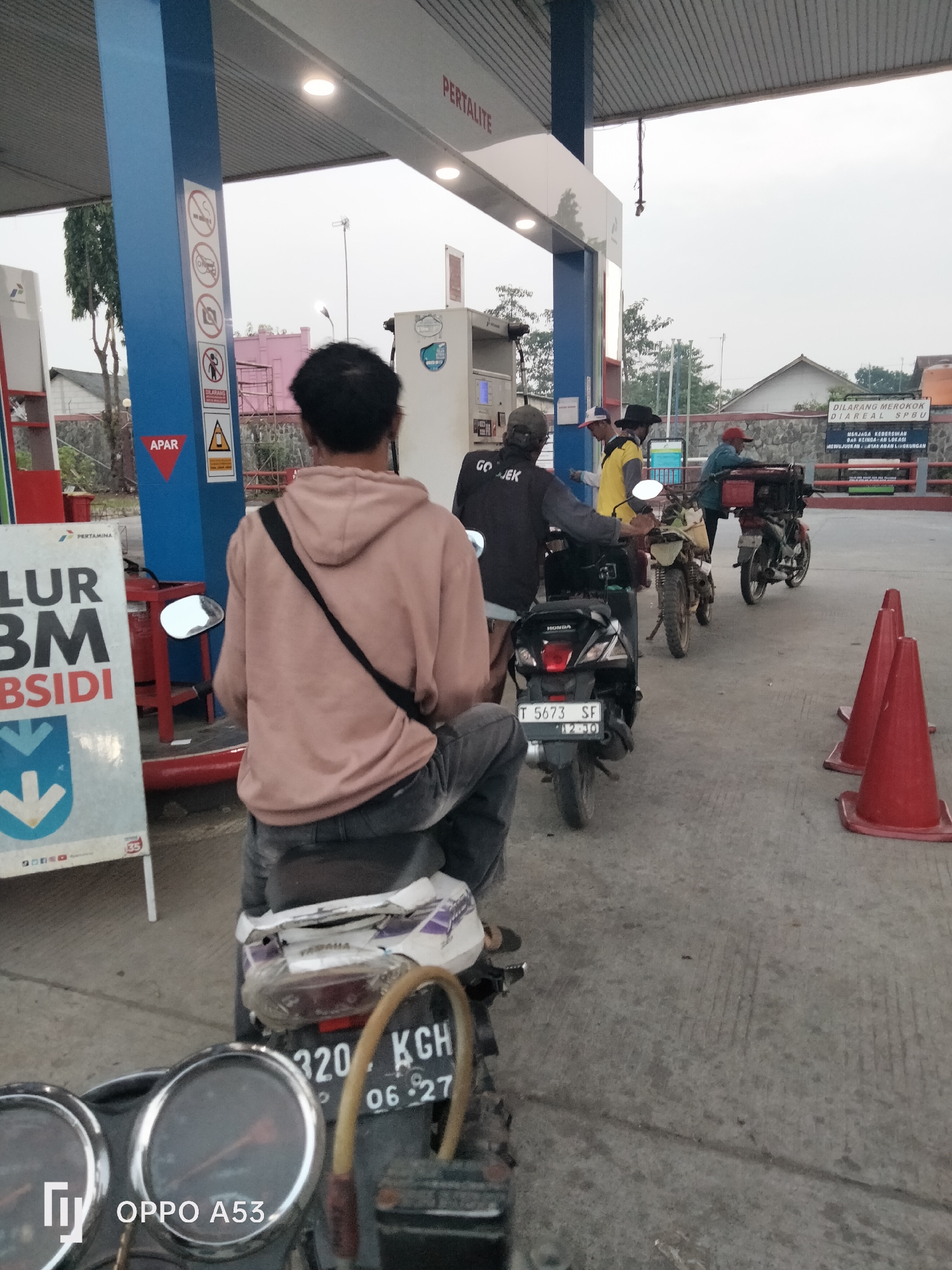 The image depicts a scene at a Pertamina gas station in Karawang, Indonesia. In the foreground, a person sits on a motorcycle, facing away from the camera. They are wearing a light pink hoodie and dark pants. The motorcycle has a black body with a white and purple sticker on the rear fender. A sign with "OPPO A53" is visible on the lower left of the motorcycle's dashboard. Behind this individual, several other motorcycles and people are lined up, presumably waiting to refuel. One person is wearing a black shirt with "GOJEK" printed on it, indicating they might be a ride-sharing driver. Another person wears a yellow shirt and a black hat.
Further back, there are gas pumps with "PERTALITE" signage. A blue sign with various symbols, including a cigarette with a slash through it, a car with a slash through it, and a person with a slash through it, is attached to one of the gas pump supports. A red triangular sign with "APAR" (Alat Pemadam Api Ringan - Portable Fire Extinguisher) is also visible on the support. In the background, there is a stone wall and some buildings. The overall atmosphere suggests a typical busy day at a gas station. The license plate of the motorcycle in the foreground is partially obscured but shows "320 KGH". Another motorcycle has a license plate that reads "T 5673 SF". Two red traffic cones are placed on the ground in the middle right of the image. The sky is overcast.
