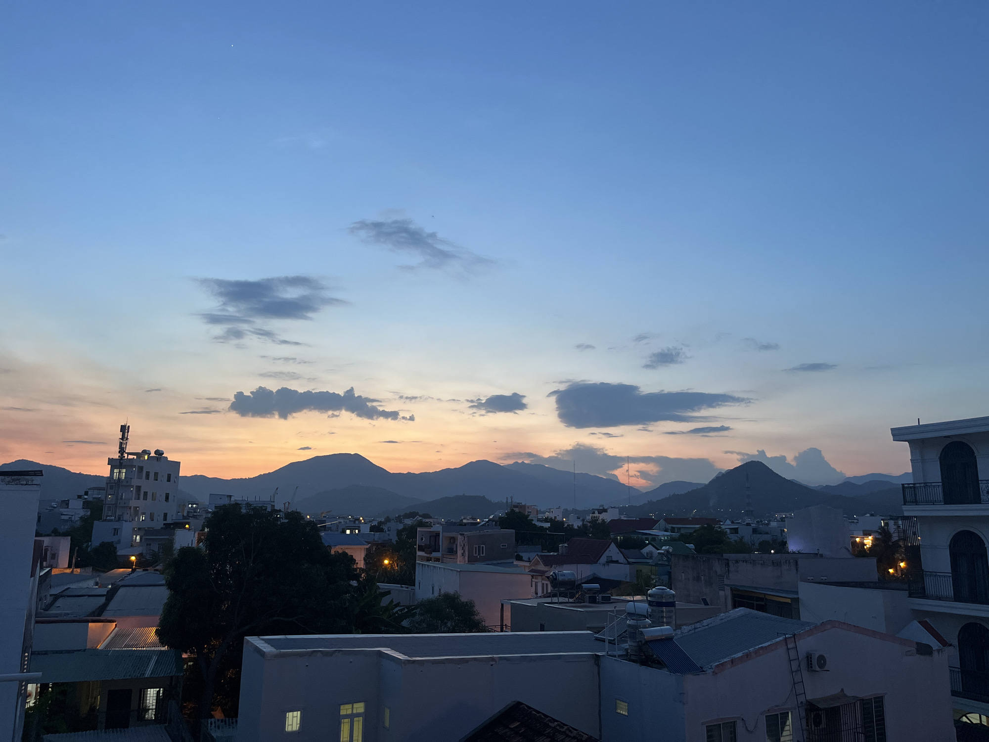 This is a wide-angle, medium-distance photograph of a city skyline at dusk, likely taken from an elevated vantage point. The sky dominates the upper half of the image, transitioning from a deep blue at the zenith to softer hues of orange and pink near the horizon, indicating the setting sun. Scattered clouds are present throughout the sky, some appearing wispy and others more defined and darker.
In the midground and foreground, a dense urban landscape is visible, characterized by a variety of buildings. These structures range from multi-story residential buildings with visible windows and balconies to lower-profile homes with corrugated metal roofs. Several buildings appear to have rooftop water tanks and air conditioning units. A prominent tall building with a communication tower on top is situated on the left side of the frame, illuminated by internal lights. Green trees, particularly a large, dark tree in the center-left, are interspersed among the buildings. The overall impression is of a lived-in urban environment.
In the background, a range of mountains forms a silhouetted horizon against the colorful sky. These mountains are dark and appear somewhat distant, with some appearing more rugged and peaked than others. The lighting suggests that it is late evening, with artificial lights starting to illuminate some of the buildings. There are no discernible people, vehicles, or animals in the frame. The atmosphere is calm and peaceful, with the natural light of the sunset blending with the emerging artificial lights of the city.