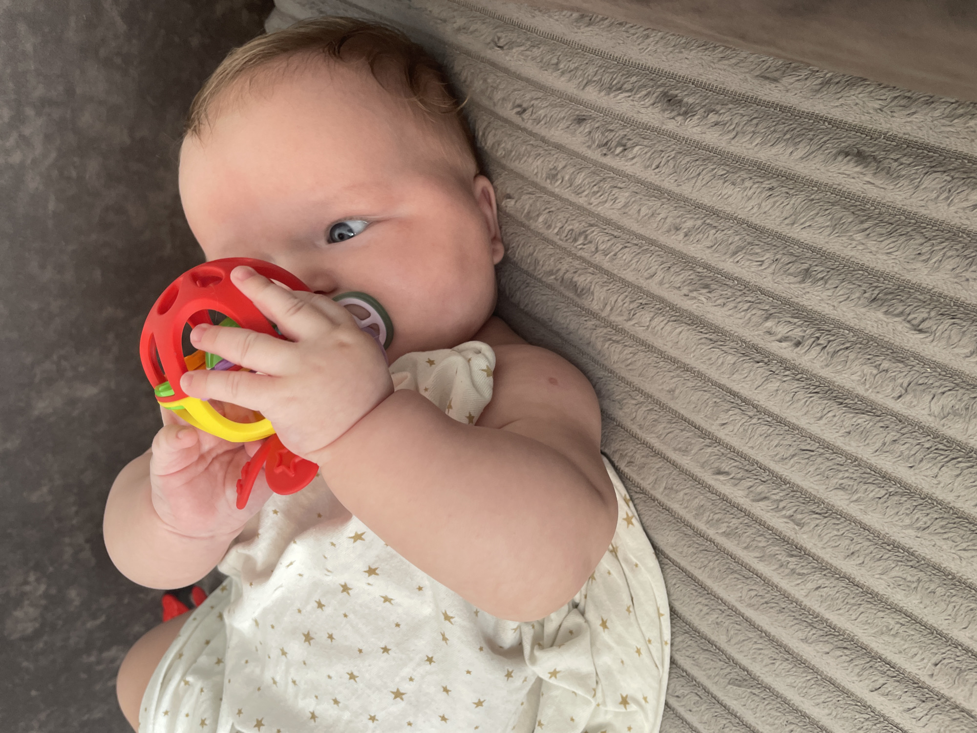 A baby is lying on a grey, textured blanket, holding a red and yellow rattle toy to their mouth. The baby is wearing a white onesie with gold star patterns. The baby's face is visible, with blue eyes and light brown hair. The background is dark and out of focus. The scene appears to be indoors.