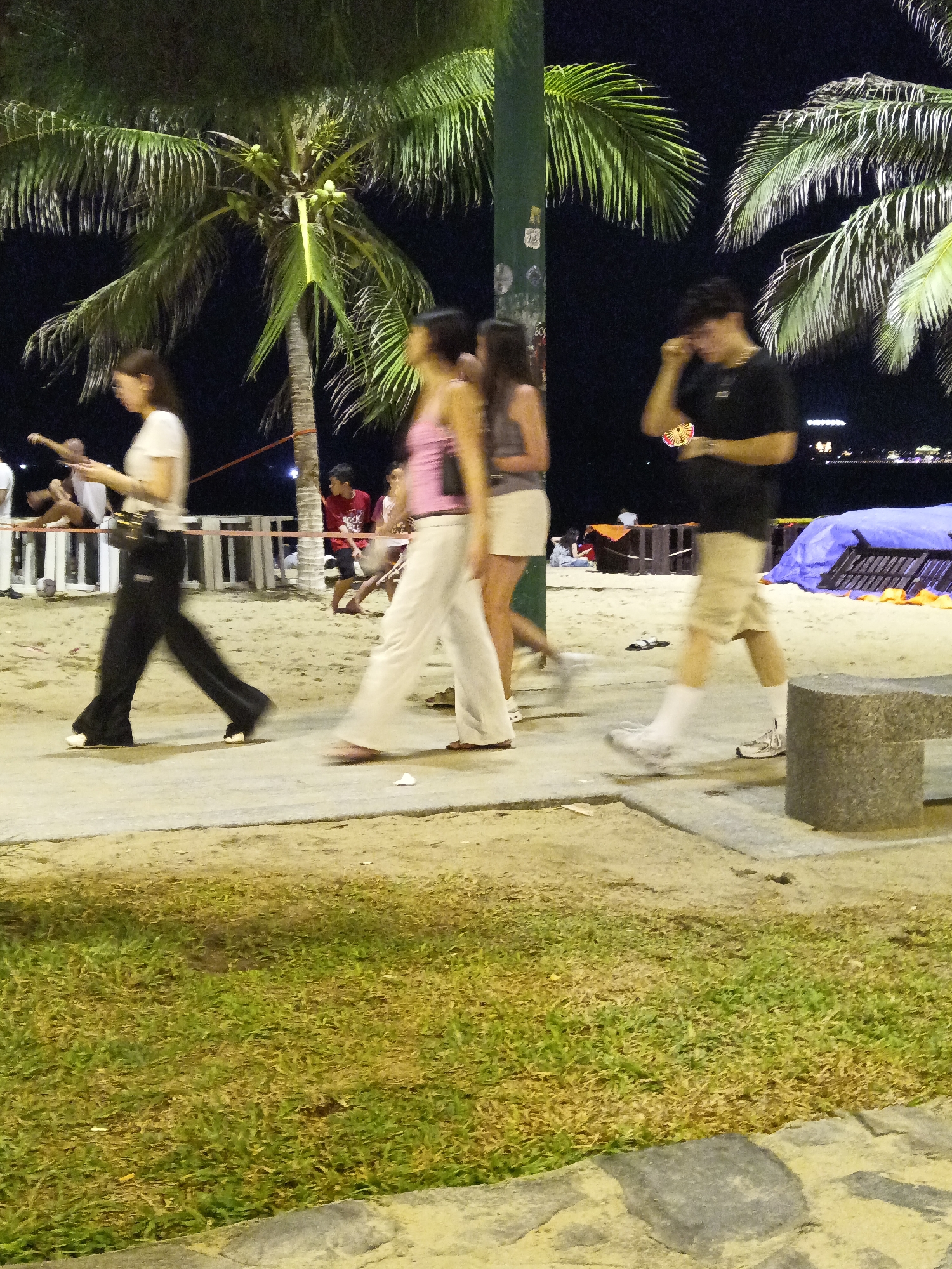 The image captures a nighttime scene on a beach promenade in Nha Trang, Vietnam, likely along Tran Phu Beach. Palm trees, characteristic of the region, are prominent, with their fronds silhouetted against the dark sky. The ground is a mix of sand and a paved walkway.
Several individuals are visible, some in motion, indicated by motion blur. On the left, a woman in a white shirt and black pants walks past, holding a mobile phone. To her right, another woman in a pink top and white pants walks away from the camera. Further to the right, a man in a black t-shirt and khaki shorts is seen talking on his phone, holding a colorful object in his hand. In the background, other people and elements of the urban beach environment are present, including what appears to be a beachside bar or restaurant, a blue tarpaulin, and beach chairs. The overall impression is of a lively evening atmosphere in a tropical coastal city.