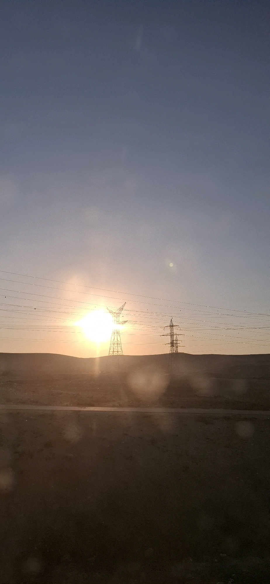 An outdoor, vertical, full shot captures the horizon during sunset in Al Sir, Israel. The sun, a bright star of yellow light, is centered just above the horizon line, partially obscured by a transmission tower. This tower is constructed of metallic latticework and stands tall amidst a desolate, hilly landscape. Another, similar transmission tower is visible to the right of the first, also silhouetted against the dusky sky. Power lines stretch horizontally across the frame, connecting the towers and disappearing into the distance. The sky is a gradient of soft blues and oranges, with a few faint circular glares of light scattered throughout. The ground is a muted brown, with subtle undulations suggesting rolling hills. A dark, paved surface, possibly a road, runs horizontally across the lower portion of the image. The overall scene conveys a sense of vastness and stillness, with the setting sun casting long shadows and creating a warm, ethereal glow.