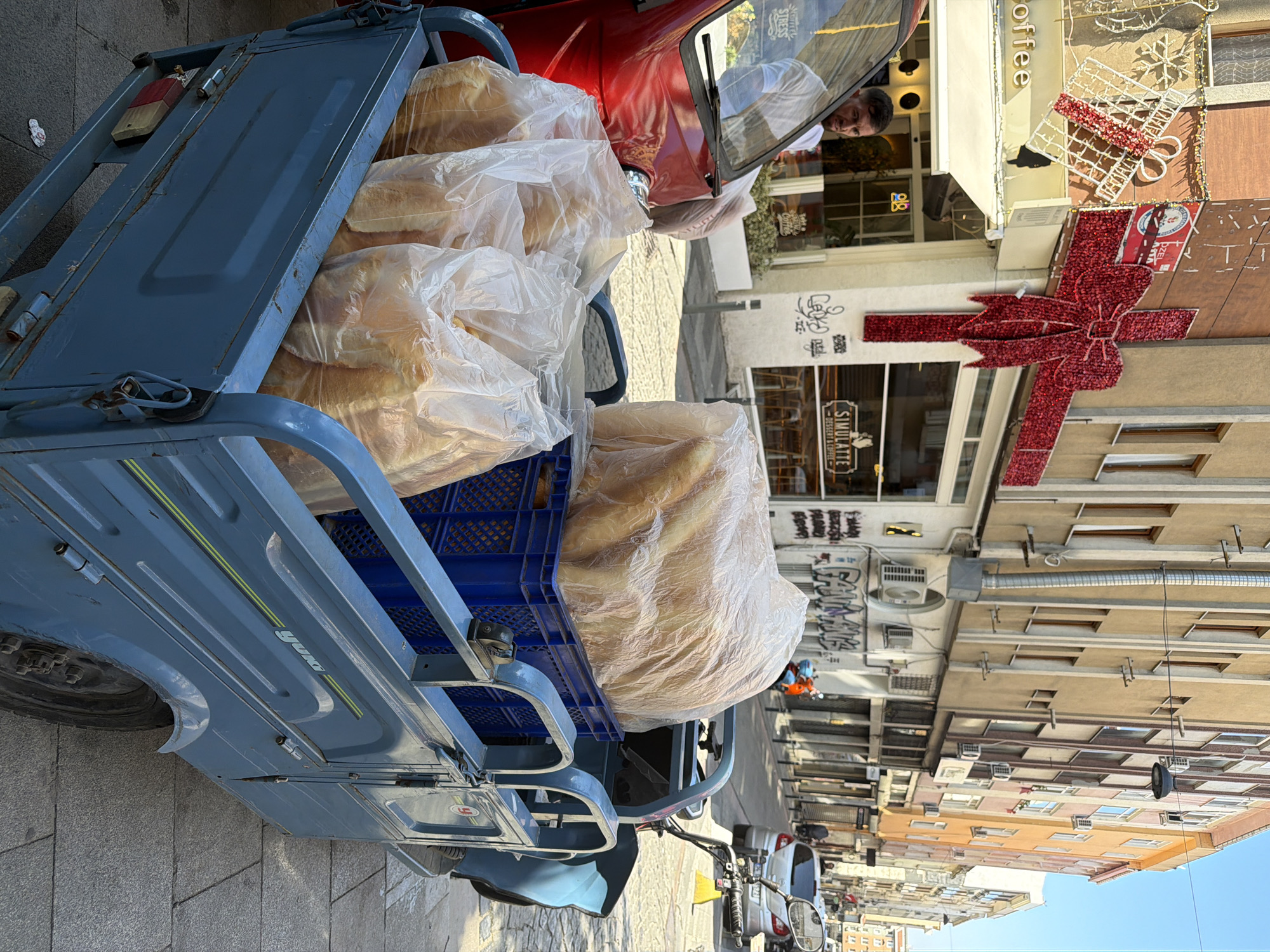 A blue three-wheeled vehicle with a cargo bed is parked on a cobblestone street in Kadıköy, Turkey. The cargo bed is filled with loaves of bread, some in clear plastic bags and others covered by a large clear plastic sheet. A red vehicle is partially visible to the left, with a man in a white shirt standing behind its windshield. In the background, buildings with storefronts line the street. Festive red ribbon decorations, resembling a large bow and gift boxes, are visible on one of the buildings. The sky is clear and blue.