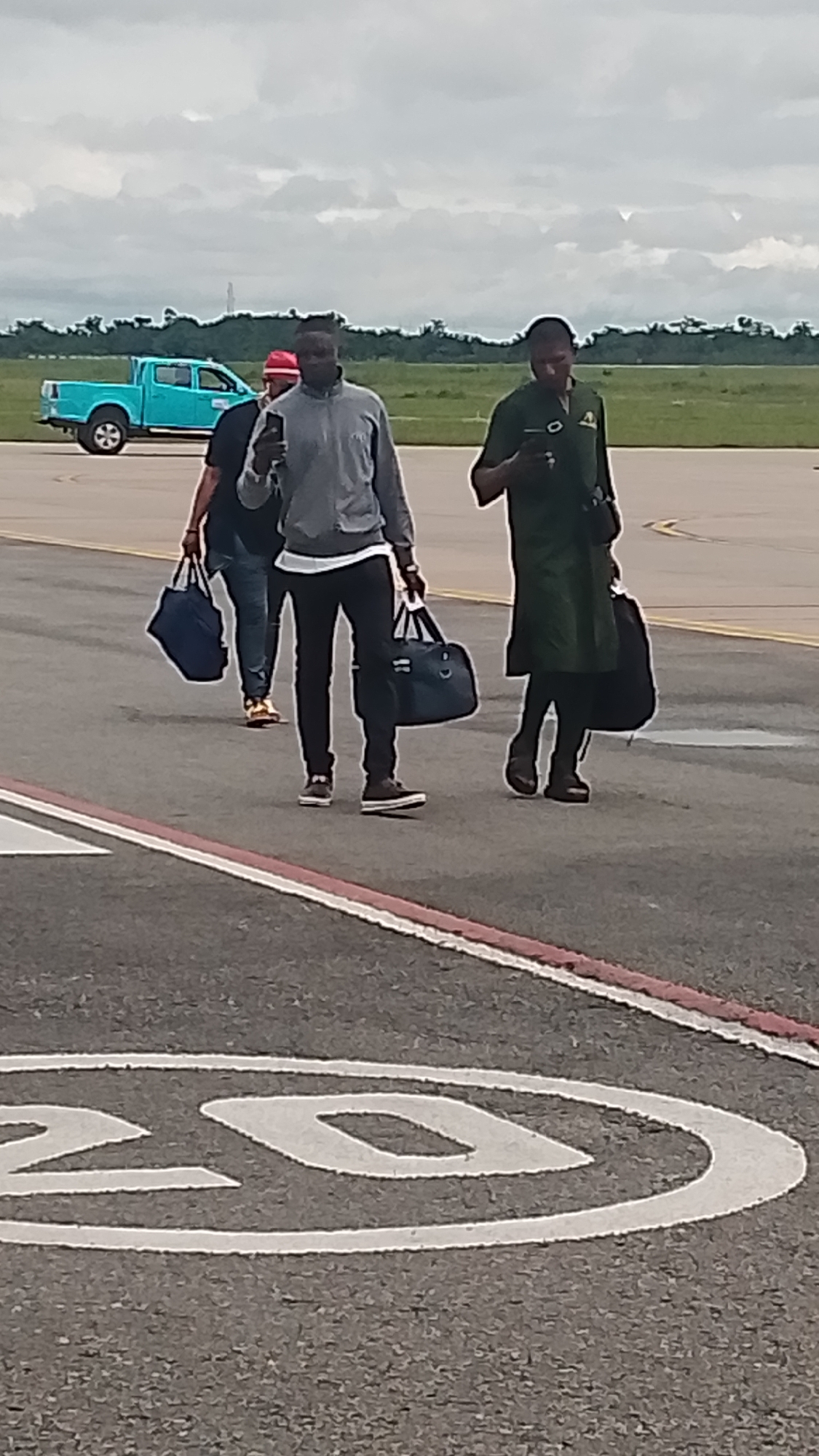 In Orerokpe, Nigeria, under a cloudy sky, three individuals are walking across an asphalt surface marked with white lines, one of which appears to be a large "20." In the background, a bright turquoise pickup truck is parked on a grassy area, with trees and a light tower visible on the horizon.
The person in the foreground, on the right, is wearing a long, dark green tunic and pants, and carrying a dark bag over their shoulder. They are looking down at their phone. To their left, a person in a gray hooded jacket and black pants is also looking at their phone, carrying a dark blue duffel bag in their left hand. Behind them, a third person, partially visible, wears a red cap and a dark shirt, carrying a dark blue bag. The overall atmosphere suggests travel or movement, possibly at an airport or a similar open transportation hub, given the presence of the truck and the vast expanse. The lighting indicates it could be daytime, with overcast conditions.